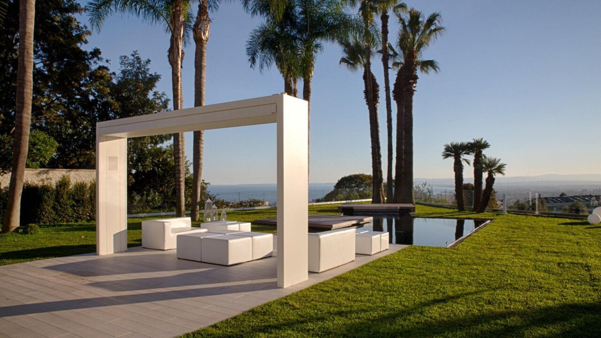 Modern outdoor lounge area with white furniture and a pergola, next to a reflecting pool and surrounded by lush green grass and tall palm trees, overlooking a scenic view of the ocean and distant hills.