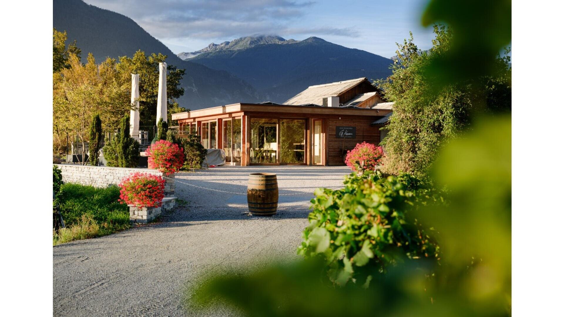 A cozy wooden building with large windows sits at the end of a gravel path, flanked by flower pots and greenery, with tall mountains and a blue sky in the background.