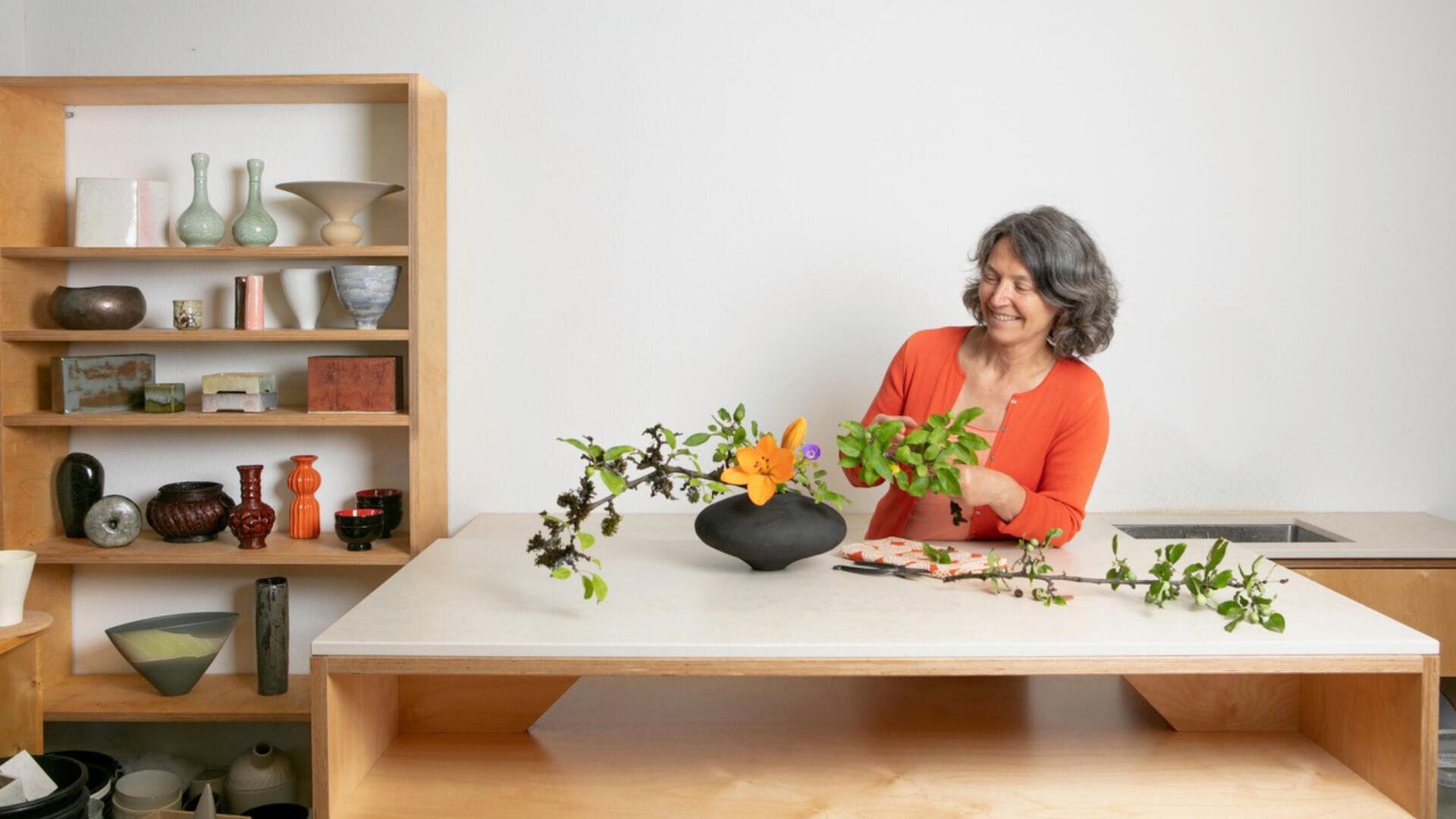 Eine Frau arrangiert lächelnd Blumen und Zweige in einer schwarzen Vase auf einem Tisch. Hinter ihr steht ein Holzregal mit Töpferwaren und Vasen, und der Raum ist einfach und modern gestaltet.