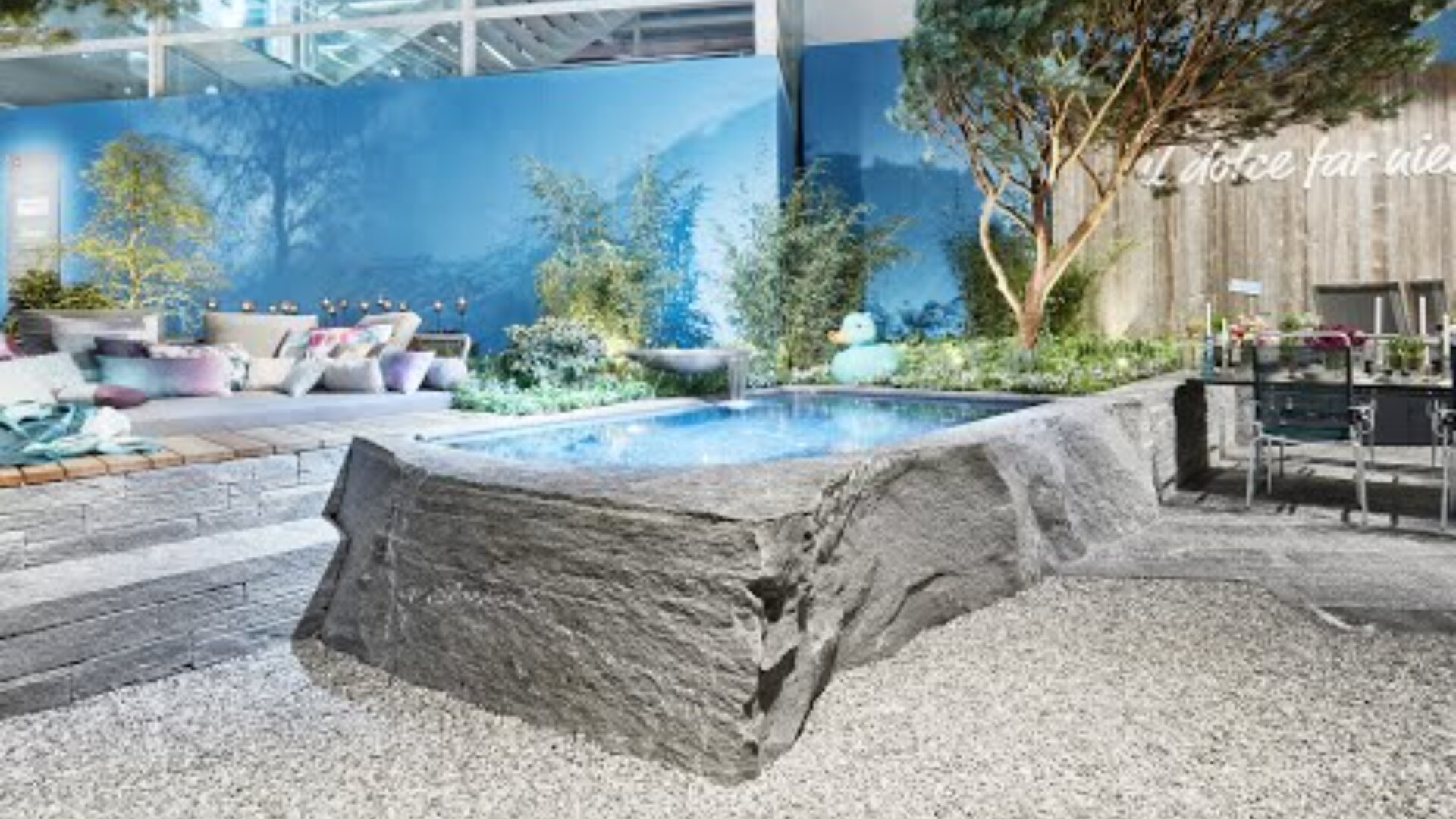 Ein moderner Garten mit einem Pool mit Steinkanten, gemütlichen Sitzgelegenheiten im Freien mit bunten Kissen, grünen Pflanzen, einem Esstisch und blauen Wänden schafft eine stilvolle und entspannende Atmosphäre.