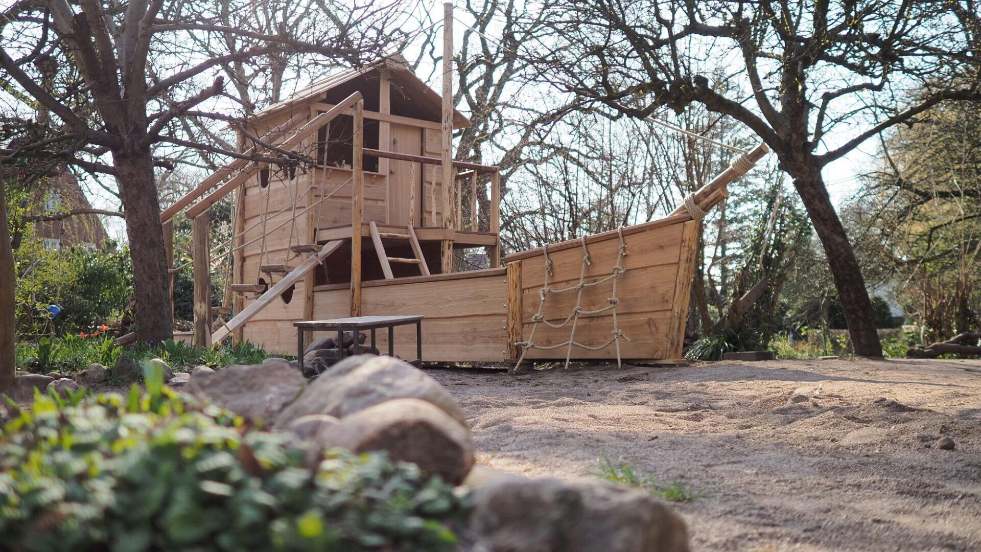 A wooden playground structure shaped like a ship, with a rope ladder, stairs, and a lookout tower, stands among trees in a sandy outdoor area. Rocks and green plants are visible in the foreground.