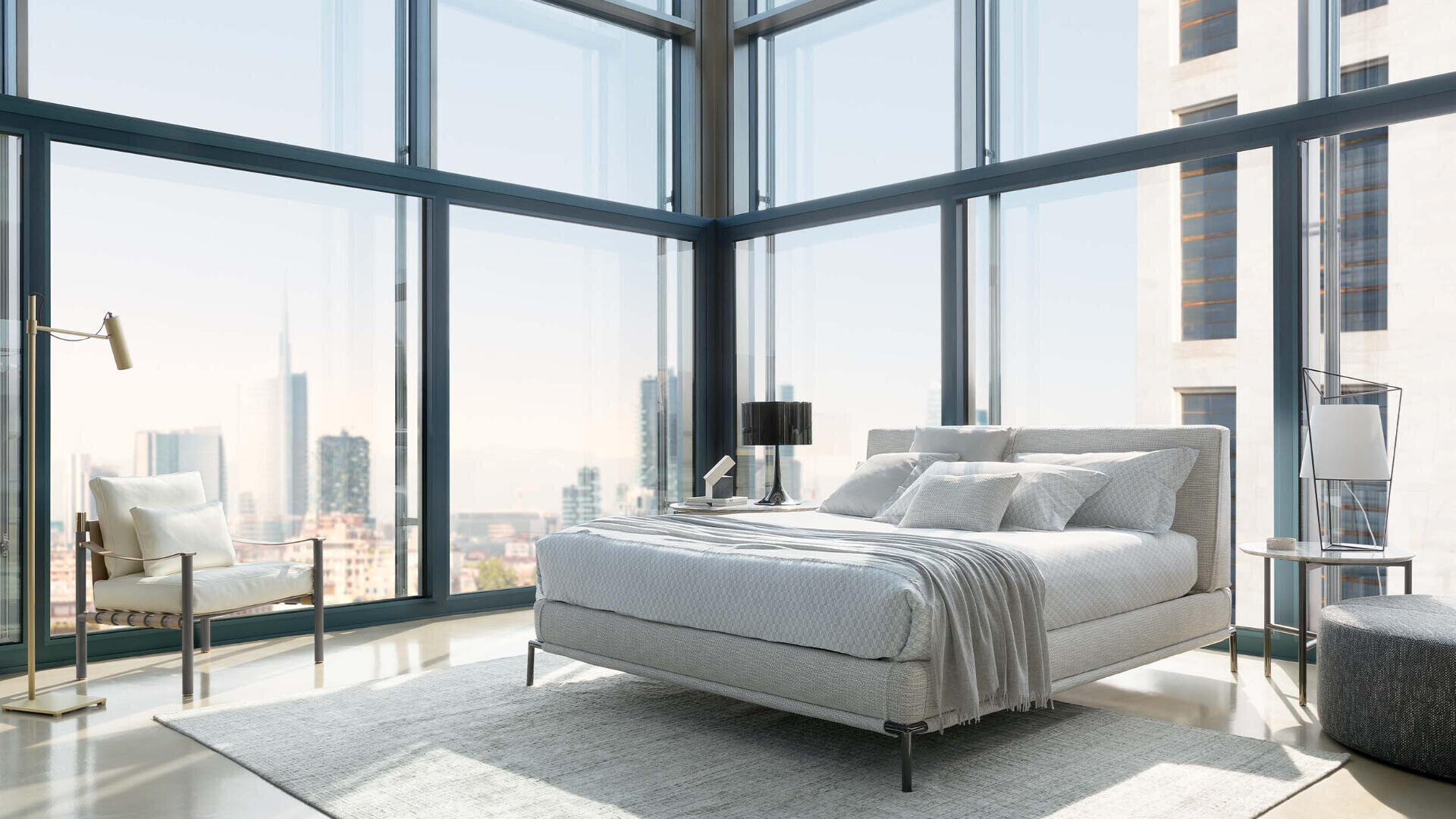 Ein modernes Schlafzimmer mit bodentiefen Fenstern und Blick auf eine Stadtlandschaft, ausgestattet mit einem grauen Bett, weißer Bettwäsche, einem grauen Sessel, einer Stehlampe, einem Beistelltisch mit Lampe und einem hellen Vorleger.