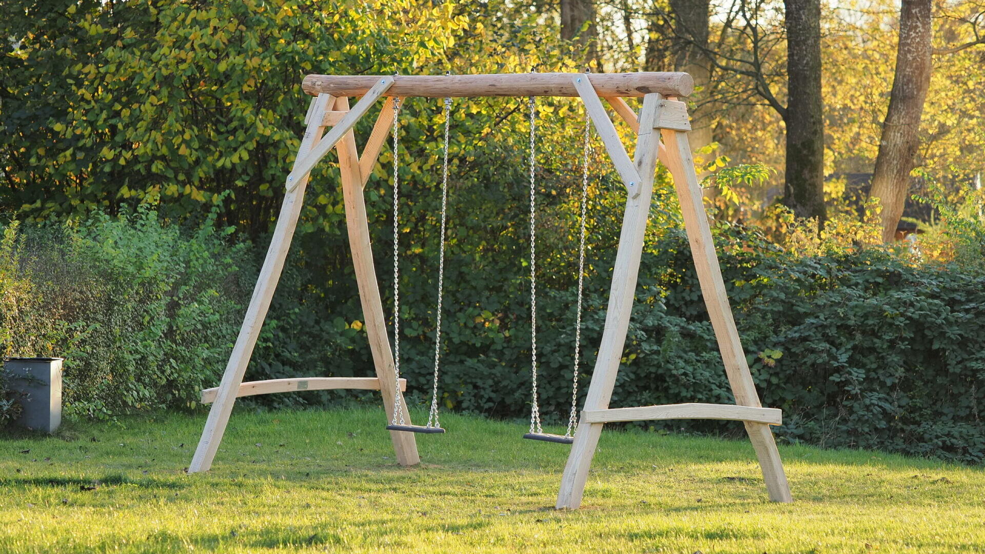 A wooden swing set with two empty swings hangs over green grass in a sunlit park, surrounded by trees and bushes.