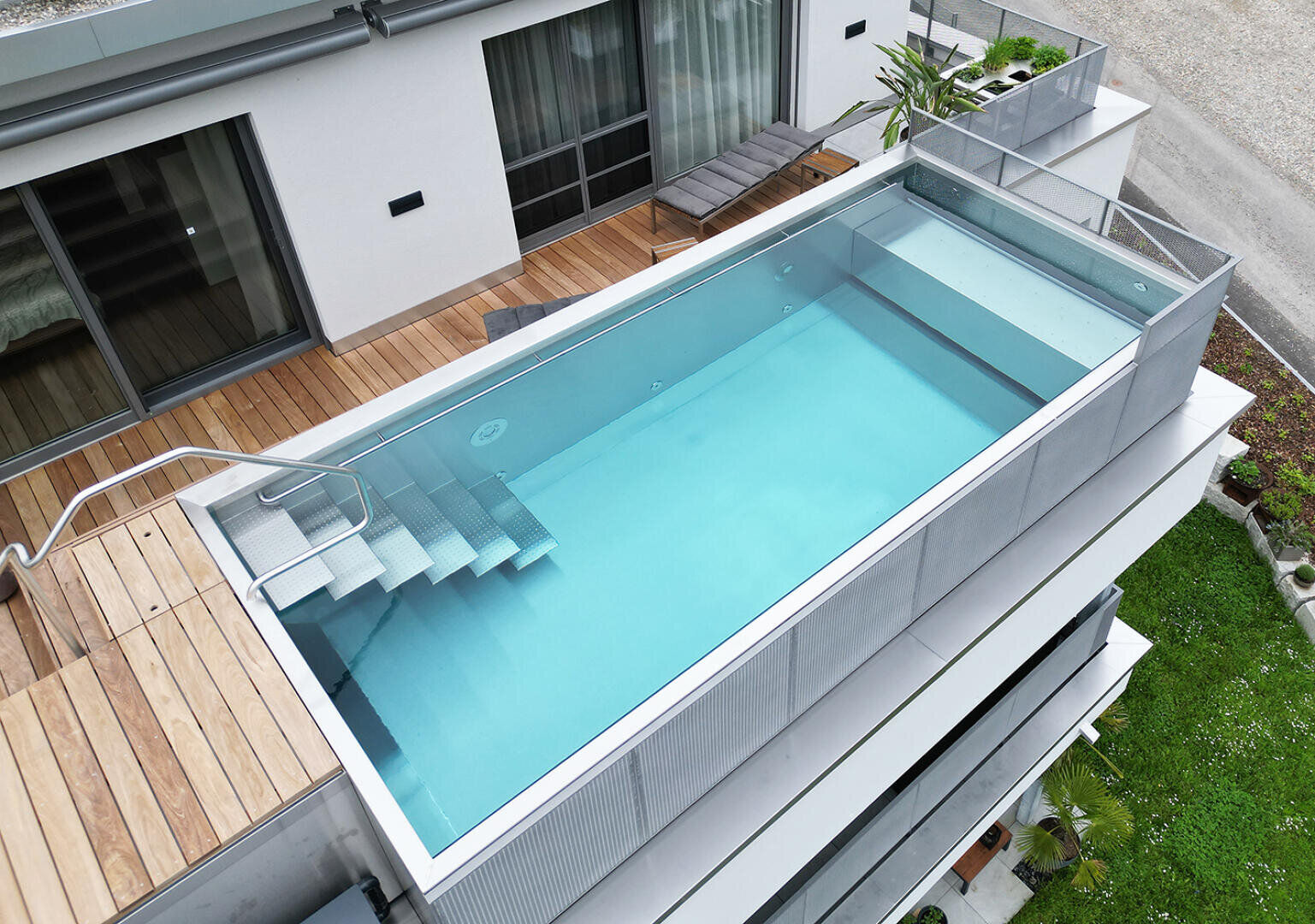 Ein modernes, rechteckiges Dachschwimmbad mit klarem, blauem Wasser und einer Metalltreppe, umgeben von einer Holzterrasse und einem Glasgeländer, das an ein modernes Wohngebäude angebaut ist.
