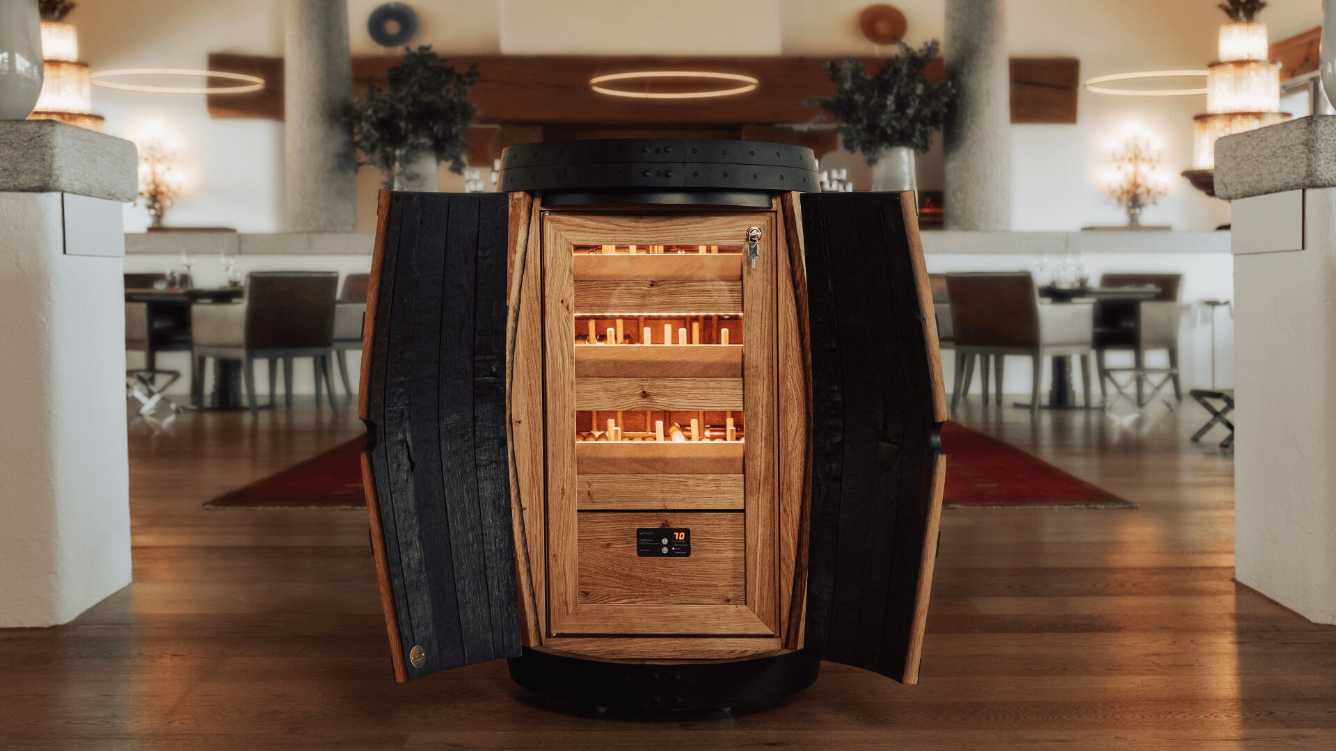 Ein Holzfass mit offenen Türen gibt den Blick auf Regale frei, die sanft beleuchtet sind und Zigarren enthalten; das Fass steht in einem modernen, eleganten Essbereich mit Tischen und Stühlen im Hintergrund.