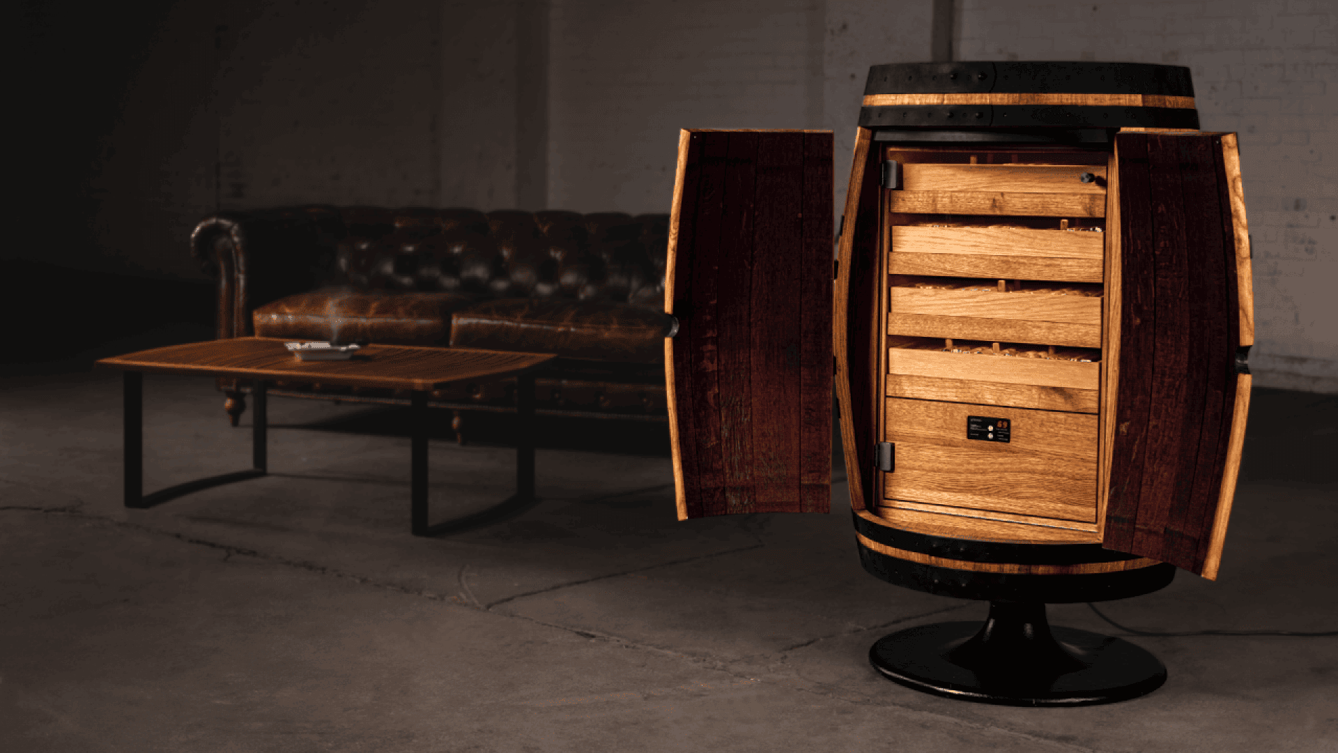 A wooden barrel repurposed as a humidor cabinet with open doors and wooden shelves stands on a pedestal in an industrial-style room with a leather sofa and wooden coffee table in the background.