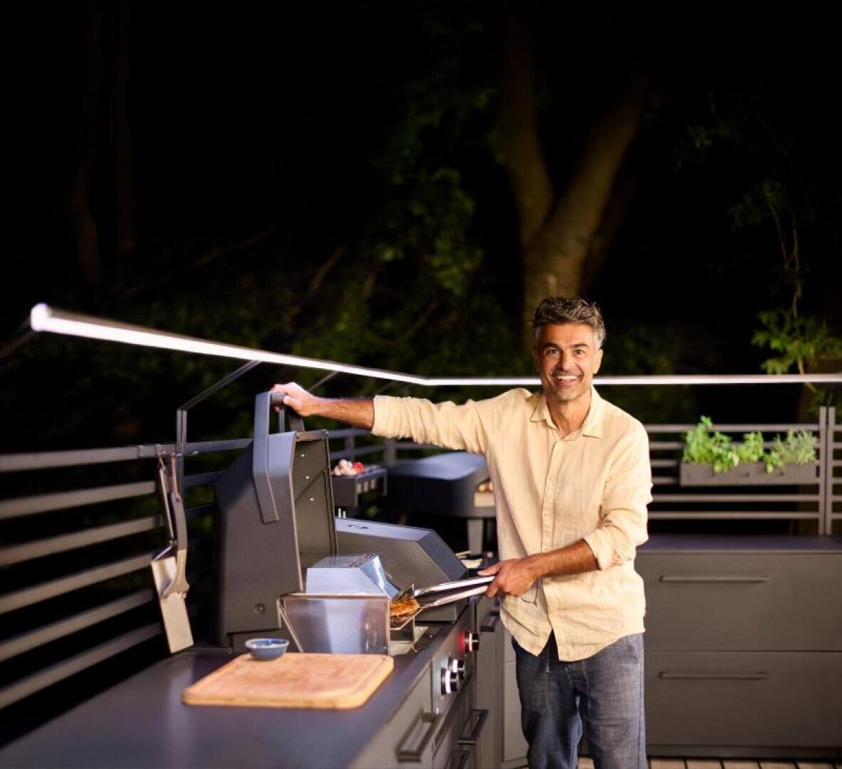 Ein Mann in einem beigen Hemd lächelt, während er nachts in einer Außenküche Essen grillt. Er hebt den Deckel eines Grills an, auf dem Tresen neben ihm liegen Utensilien und ein Holzschneidebrett. Im Hintergrund sind Bäume und Pflanzen zu sehen.