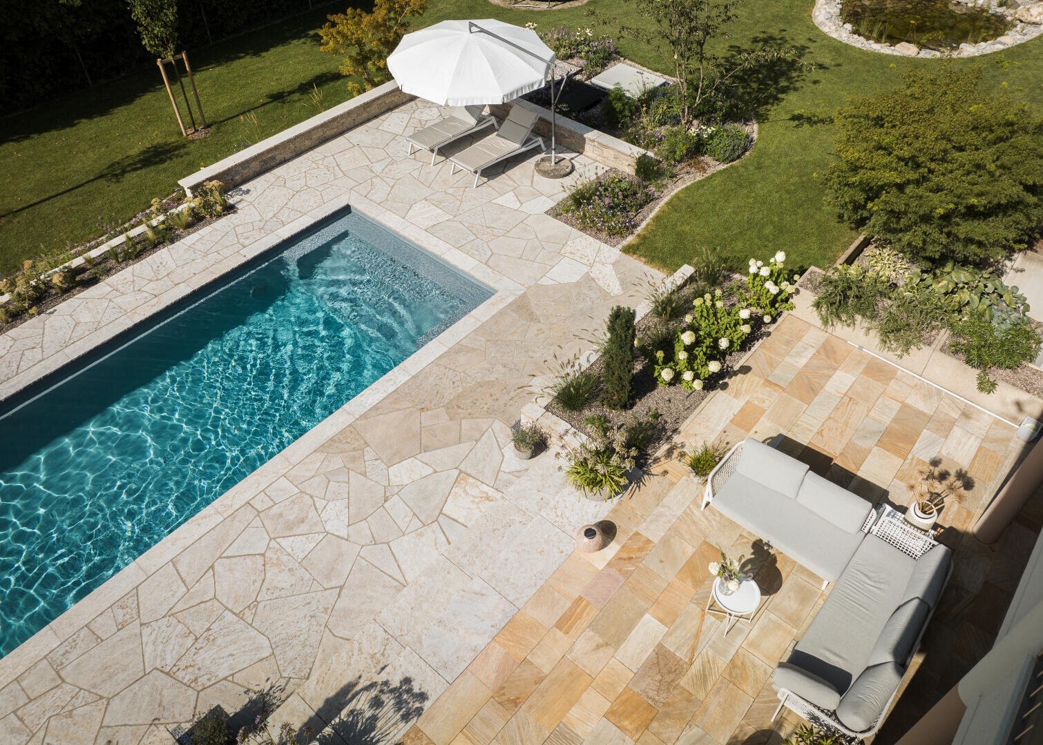 Luftaufnahme eines Hinterhofs mit einem rechteckigen Swimmingpool, einer Steinterrasse, einem Sofa, einem Tisch, Topfpflanzen, zwei Liegestühlen und einem weißen Sonnenschirm, umgeben von grünem Rasen und Bäumen.