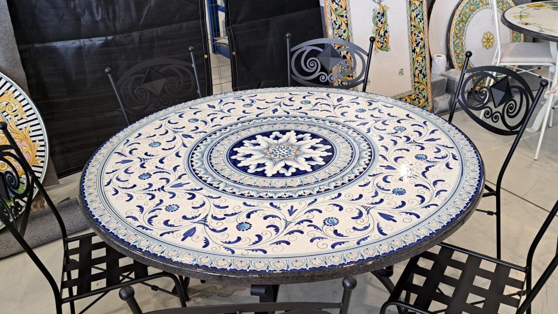 A round table with an ornate blue and white ceramic mosaic top featuring intricate floral and geometric patterns, surrounded by four black metal chairs with decorative backs.