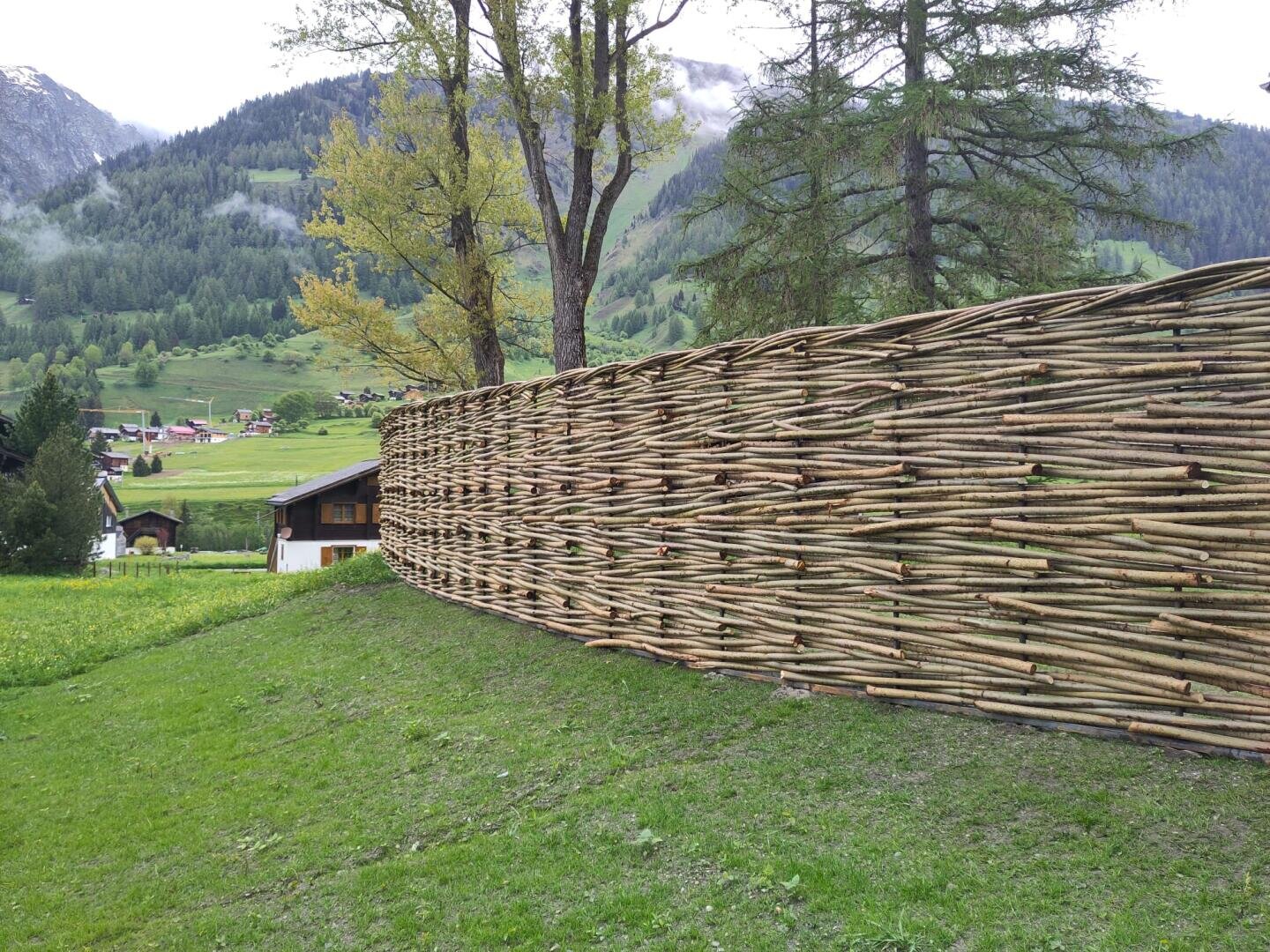 Ein hoher geflochtener Zaun aus dünnen Holzästen steht auf einem grasbewachsenen Hang, mit Bäumen, Hügeln und einem kleinen Haus im Hintergrund unter einem bewölkten Himmel.