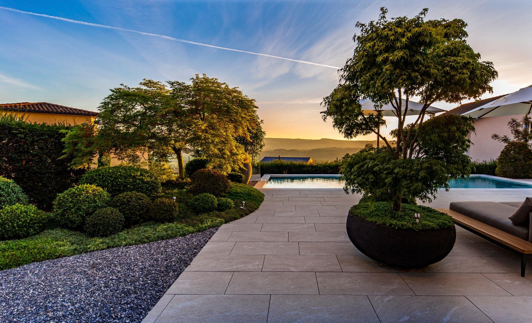 A garden at sunset with a tree in a large planter, well-tended shrubs, flagstones, gravel and a pool with sun loungers, overlooking distant hills under a clear sky.