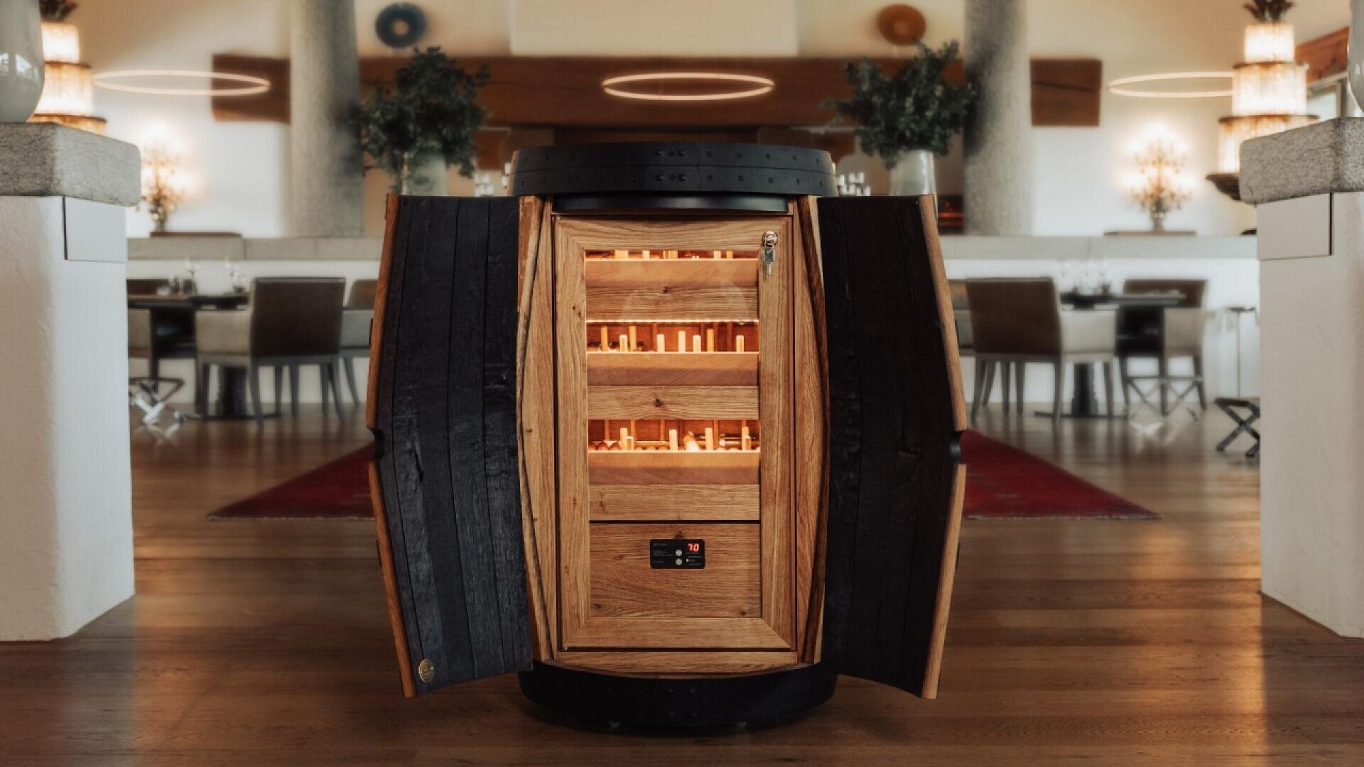 A wooden wine barrel, converted into a wine cabinet with glowing shelves, stands open in an upscale restaurant with wooden floors, set tables, and elegant decor in the background.