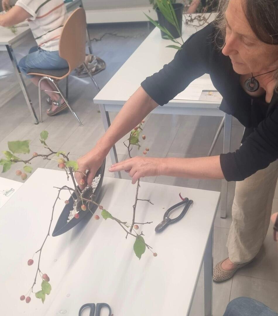 Eine Frau arrangiert einen Zweig mit Blättern und kleinen Beeren in einer schwarzen Vase auf einem weißen Tisch. Scheren und Blumenwerkzeuge liegen in der Nähe. Andere Personen arbeiten an separaten Tischen im Hintergrund.