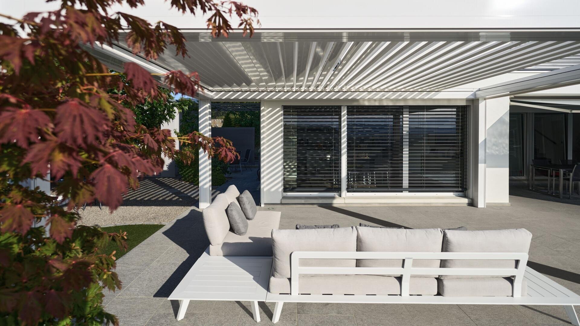 A modern outdoor patio features a white sofa with light gray cushions under a slatted pergola. A red-leaved tree is in the foreground, and large windows with blinds reflect the sun in the background.