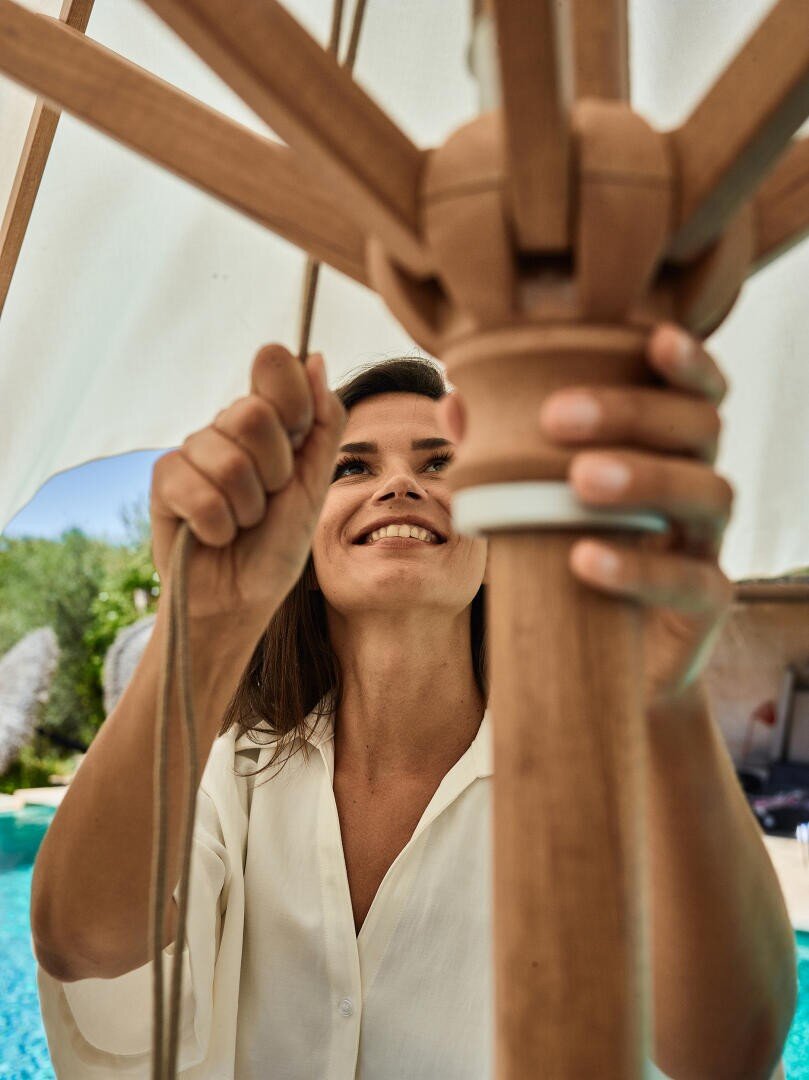 Eine Frau in einem weißen Hemd lächelt, während sie einen großen hölzernen Terrassenschirm an einem Pool aufspannt oder ausrichtet, mit Grünpflanzen und einer sonnenbeschienenen Umgebung im Hintergrund.