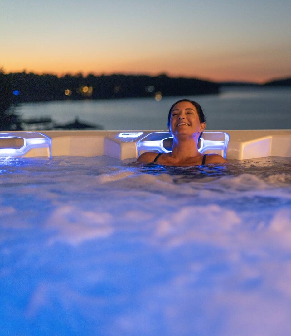 A person relaxes in a whirlpool with blue lights, eyes closed and smiling, while the sun sets over a calm lake and silhouetted trees in the background.