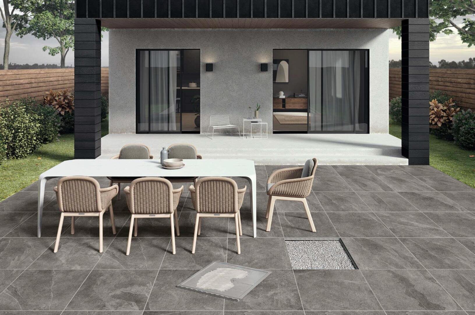 Eine moderne Außenterrasse mit grauen Steinfliesen, einem weißen Tisch, sechs Korbsesseln und Glasschiebetüren, die zu einem modernen Haus führen. Der Bereich ist sauber und von einem Holzzaun und Landschaftsbau umgeben.