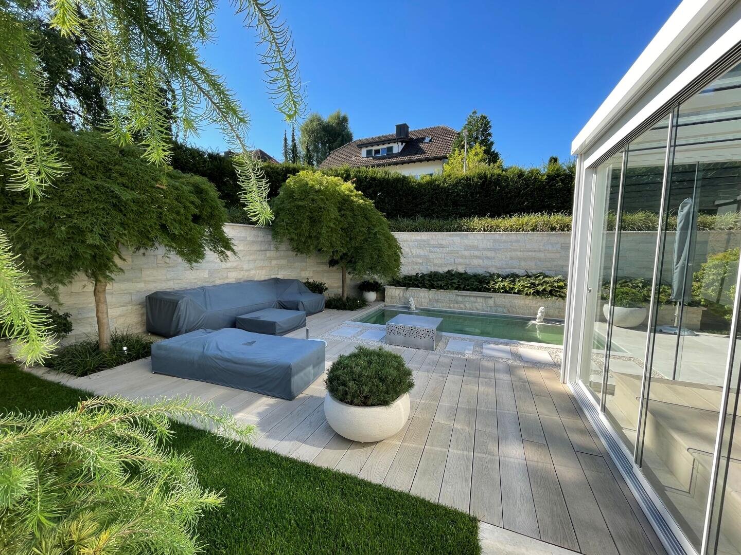 Ein moderner Hinterhof mit überdachten Gartenmöbeln, einem kleinen Pool, Topfpflanzen, grünem Rasen und einer weißen Steinmauer, umgeben von Bäumen und Sträuchern unter einem klaren blauen Himmel.