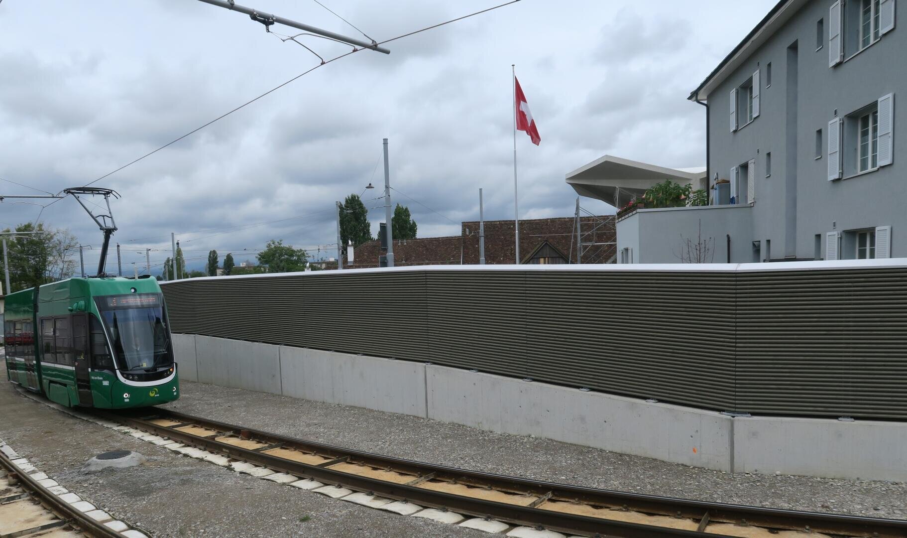 Ein grünes Tram fährt auf gekrümmten Gleisen neben einer hohen grauen Schallschutzwand, eine Schweizer Fahne weht neben modernen Gebäuden unter einem bewölkten Himmel.