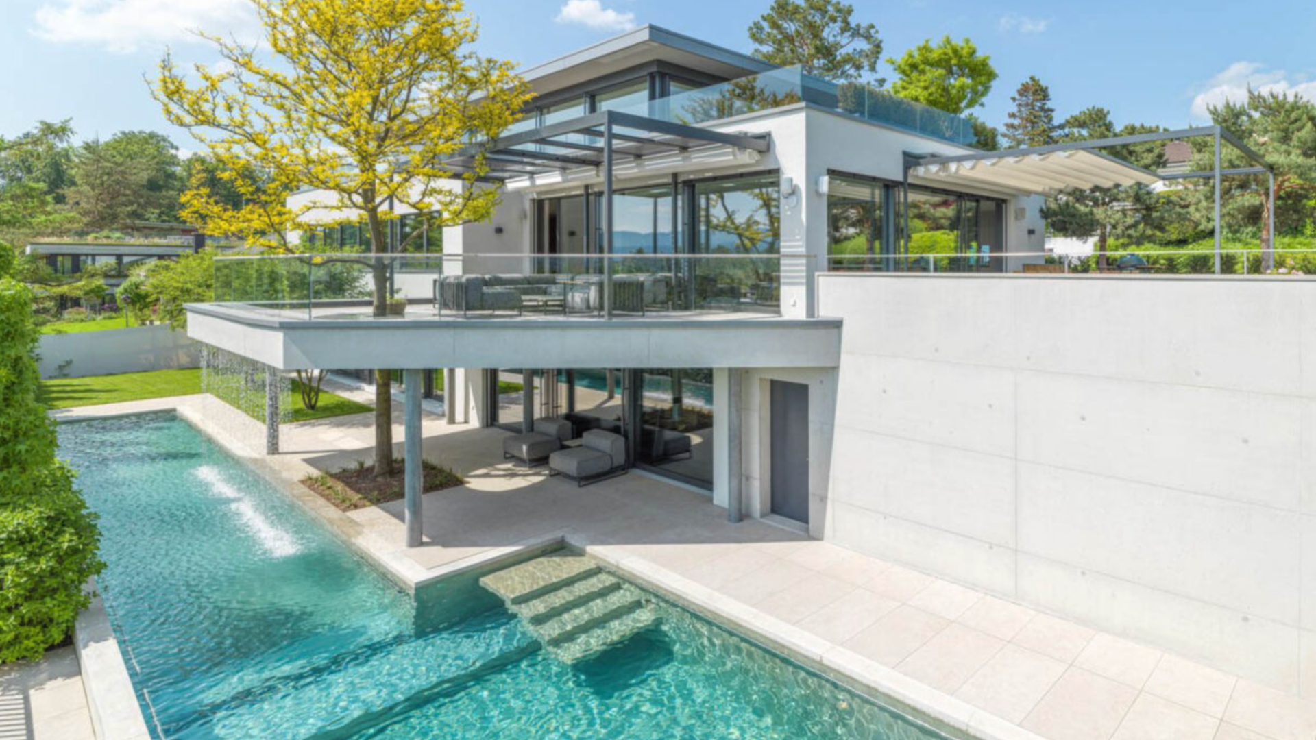 Moderne weiße Villa mit großen Glasfenstern, Außenterrasse und Sitzecke mit Blick auf einen Swimmingpool im Grünen unter einem klaren blauen Himmel.
