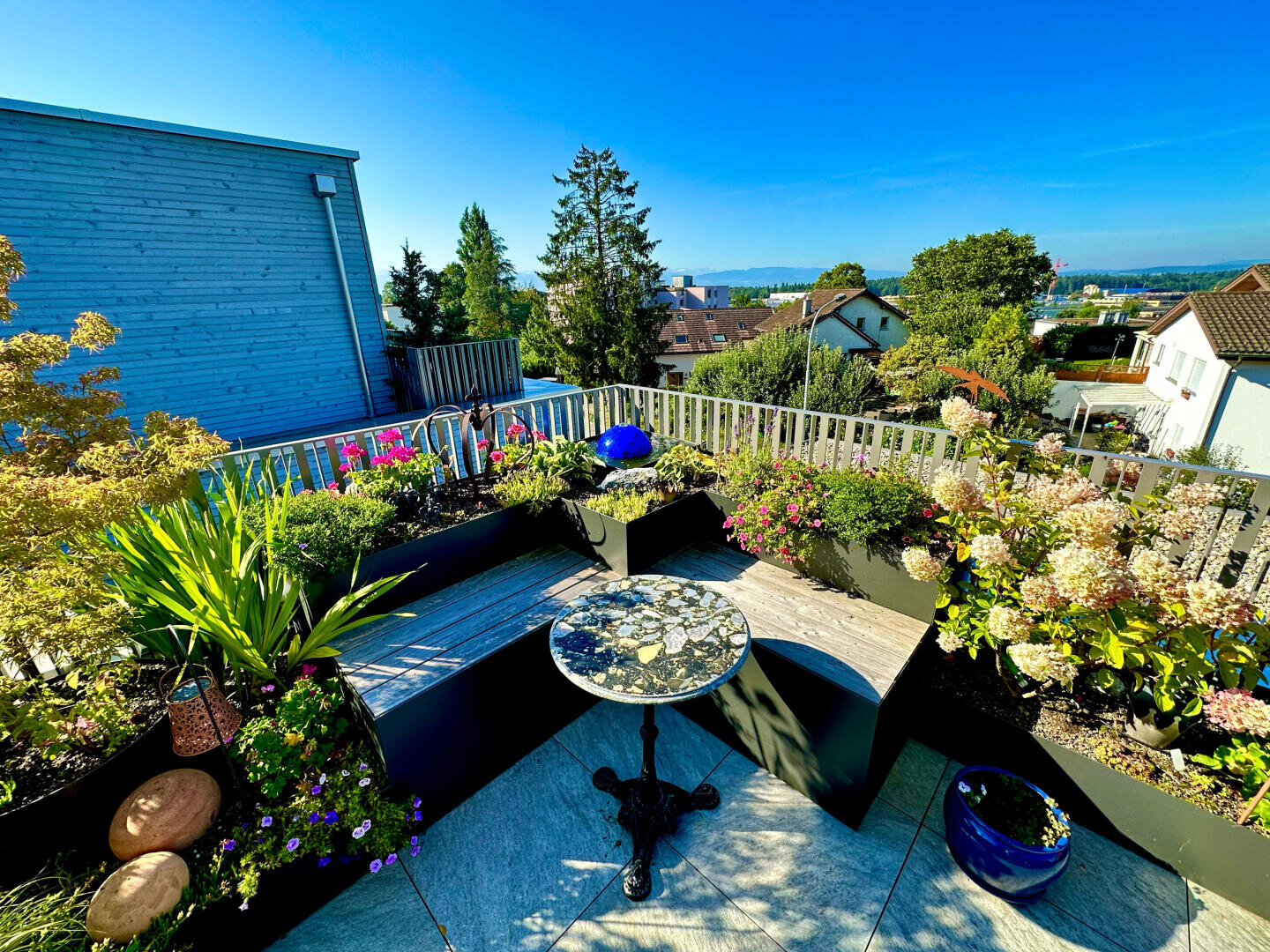 Ein sonniger Dachgarten mit einem runden Tisch aus Mosaik, geschwungenen Holzbänken und Pflanzgefäßen mit bunten Blumen und Grünpflanzen. Im Hintergrund sind Häuser und Bäume unter einem klaren blauen Himmel zu sehen.