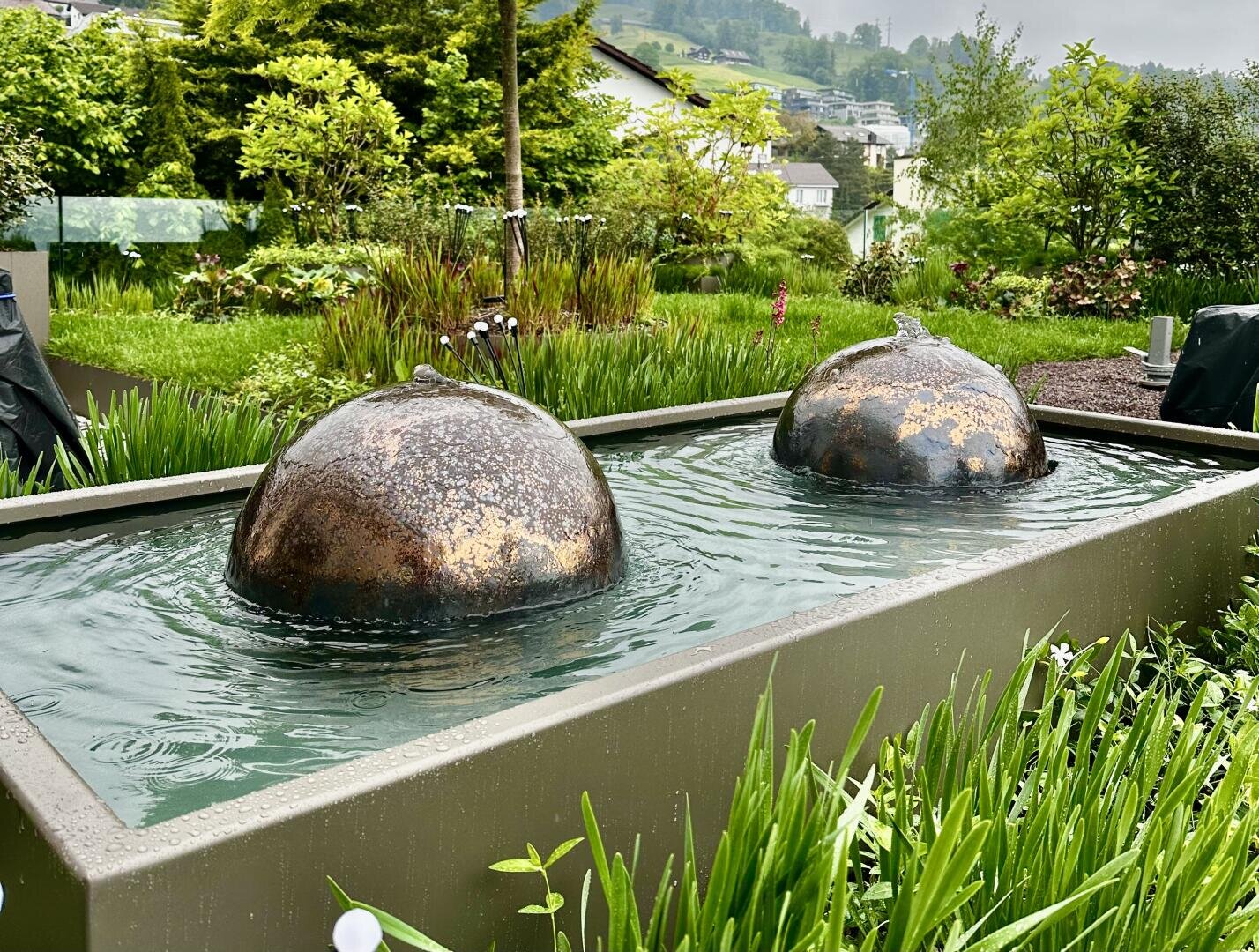 Zwei kuppelförmige Metallbrunnen sprudeln sanft in einem rechteckigen Teich, der von üppigen grünen Pflanzen und Bäumen in einem angelegten Garten umgeben ist.