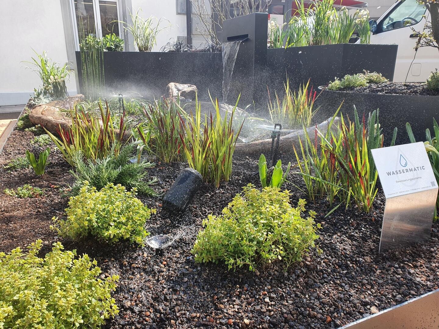 Ein angelegter Garten mit grünen und roten Zierpflanzen wird mit Sprinklern bewässert. In der Erde vor den Pflanzen befinden sich ein kleines Wasserspiel und ein Schild mit der Aufschrift WASSERMATIC.