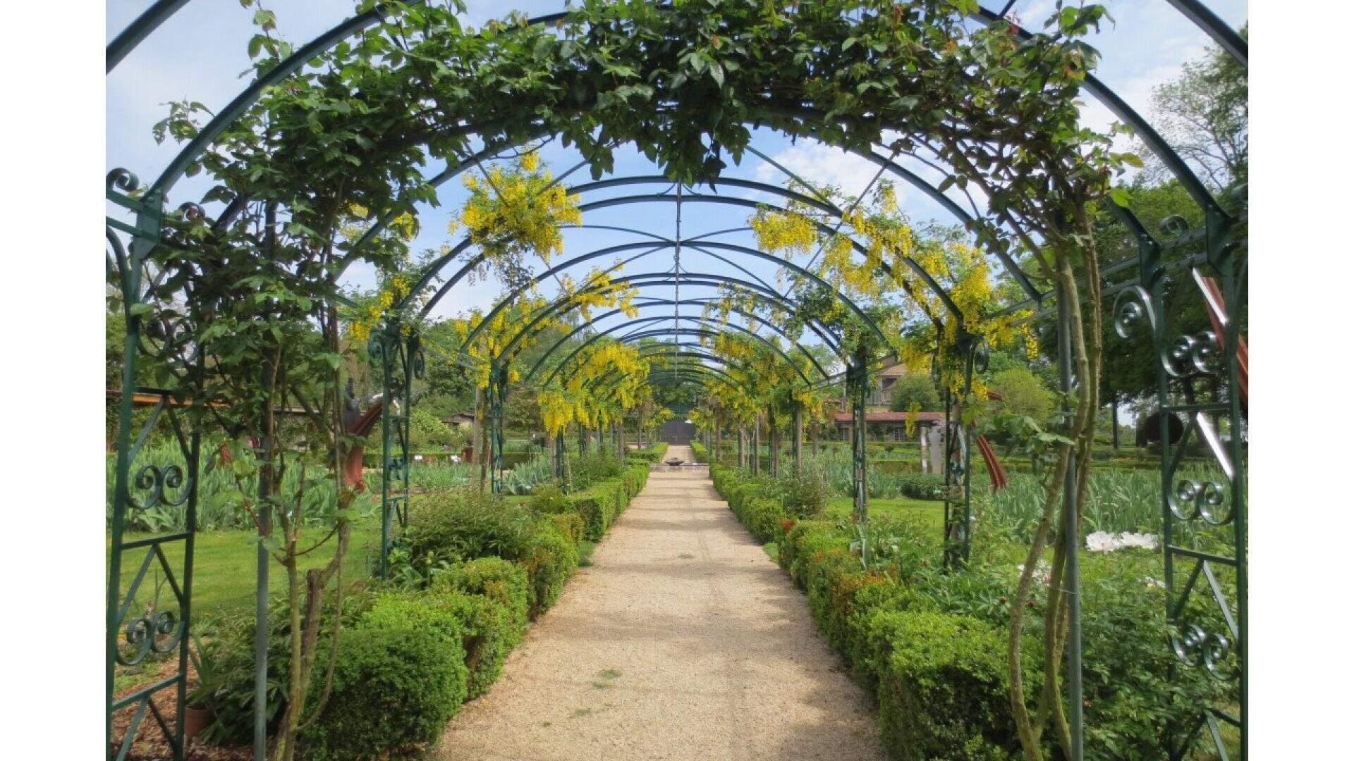 A gravel path runs under a series of metal arches covered with green climbing plants, surrounded by neatly trimmed bushes and lush gardens on a bright, partly cloudy day.