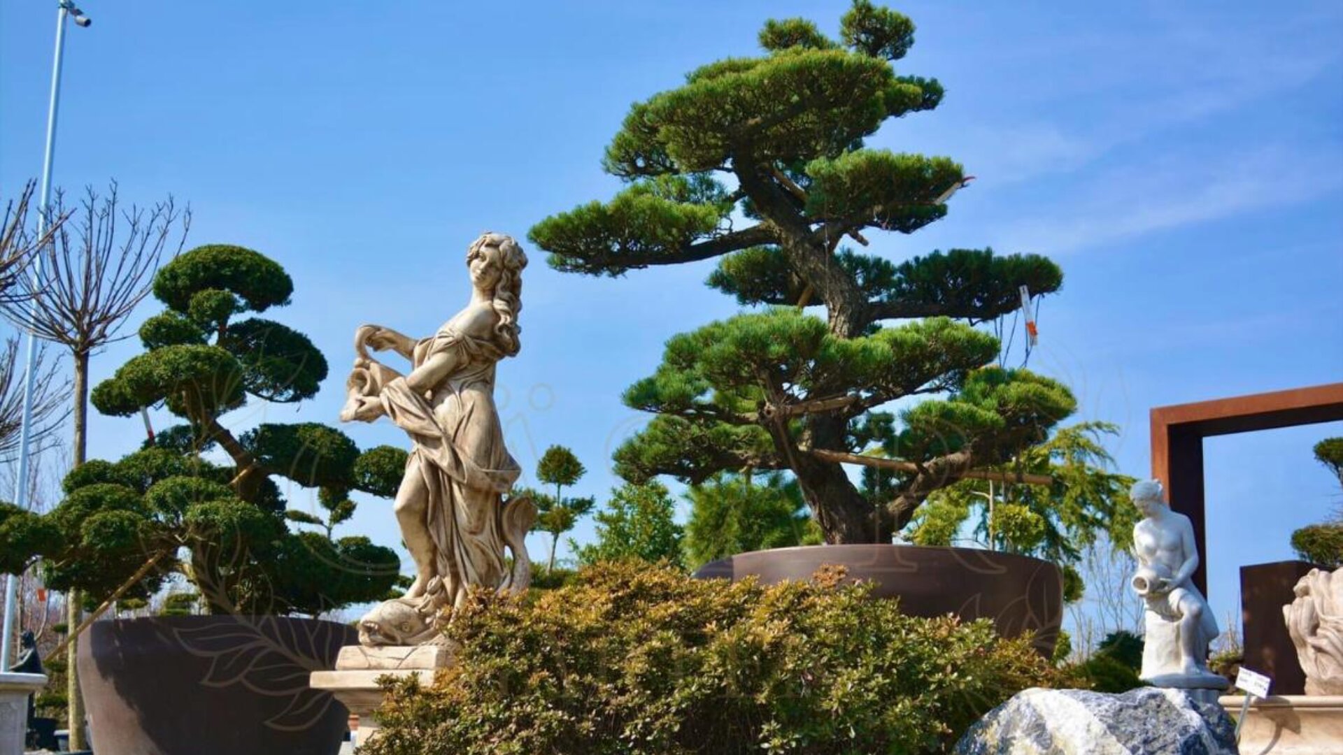 A marble statue of a woman stands in a landscaped garden with neatly trimmed trees, shrubs, and a bright blue sky in the background. Another smaller statue is visible to the right.
