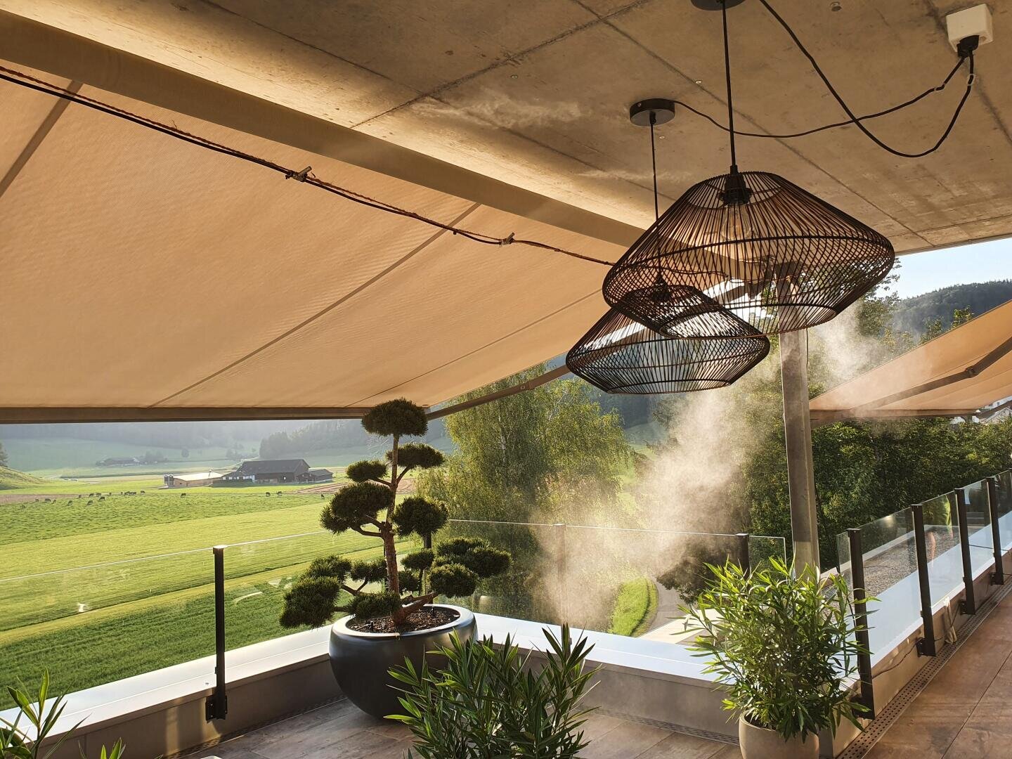 Eine moderne Terrasse mit Hängelampen, Topfpflanzen und Sonnensegeln bietet einen Blick auf grüne Felder und Bäume. Das Sonnenlicht dringt ein, während Nebel aus einer Leuchte aufsteigt und eine ruhige Atmosphäre im Freien schafft.