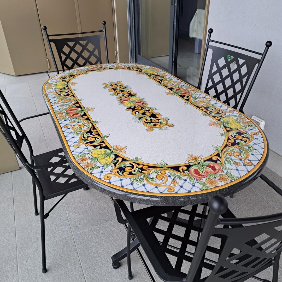 Ein dekorativer ovaler Tisch mit einem farbenfrohen, verschnörkelten Blumen- und Schneckenmuster auf der Tischplatte, umgeben von vier schwarzen Metallstühlen mit vergitterten Rückenlehnen, die auf einem hellen Fliesenboden in einem Außen- oder Terrassenbereich stehen.