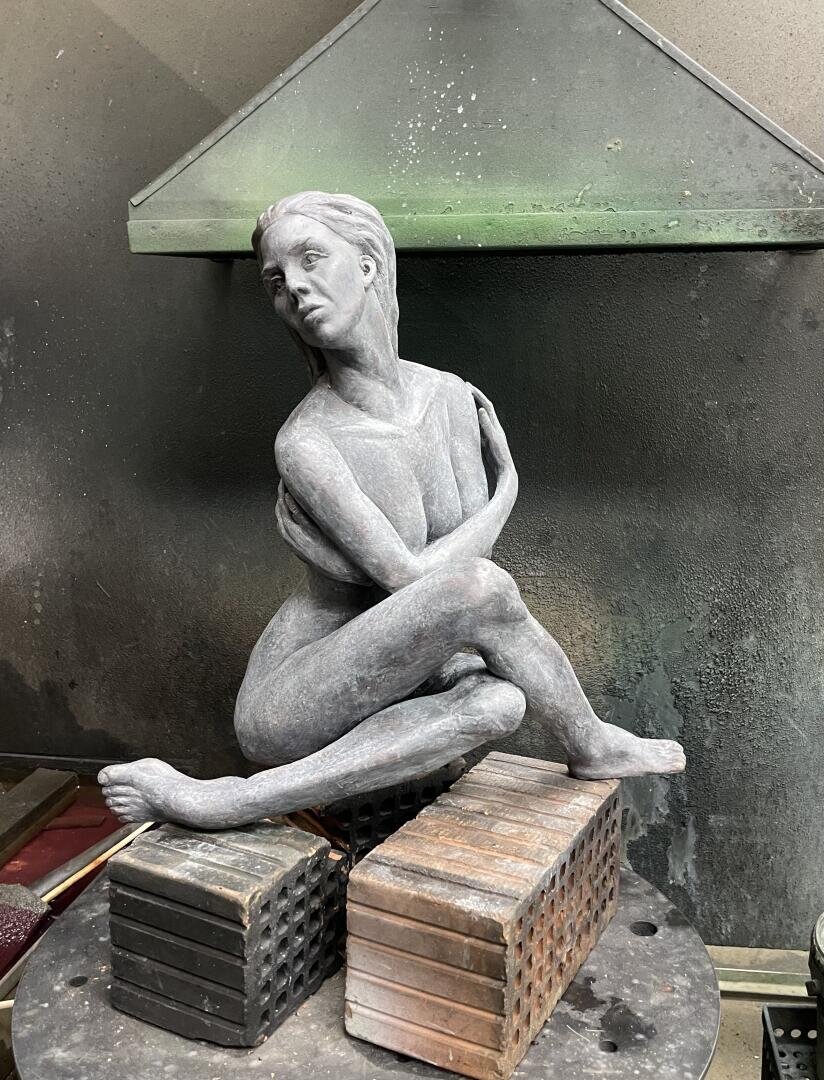 Eine Steinskulptur einer sitzenden nackten Frau mit gekreuzten Beinen und vor der Brust verschränkten Armen, die auf zwei gestapelten Metallblöcken in einer industriellen Werkstatt steht.