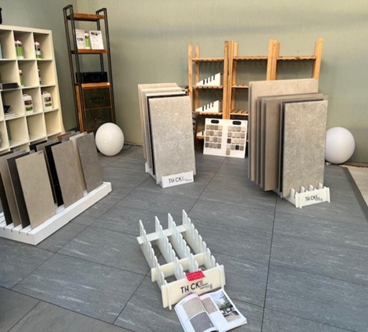 Ausstellungsbereich mit mehreren Ständern mit großen Fliesenmustern in Grau- und Beigetönen, Regalen mit Broschüren und Fliesenmustern sowie einigen Dekorationsartikeln auf einem gefliesten Boden.