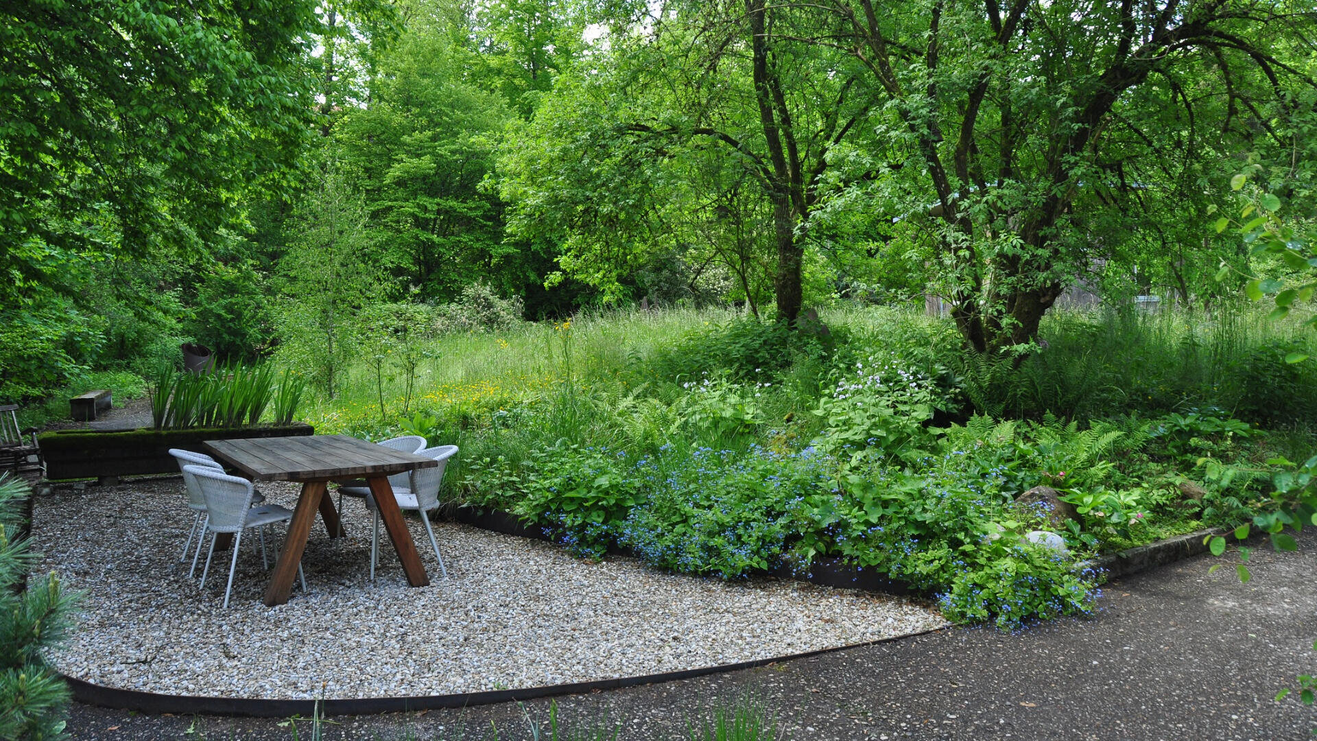 Ein kleiner Sitzbereich im Freien mit einem Holztisch und vier weißen Stühlen auf Kies, umgeben von üppig grünen Bäumen, Sträuchern und Wildblumen in einer natürlichen Gartenumgebung.