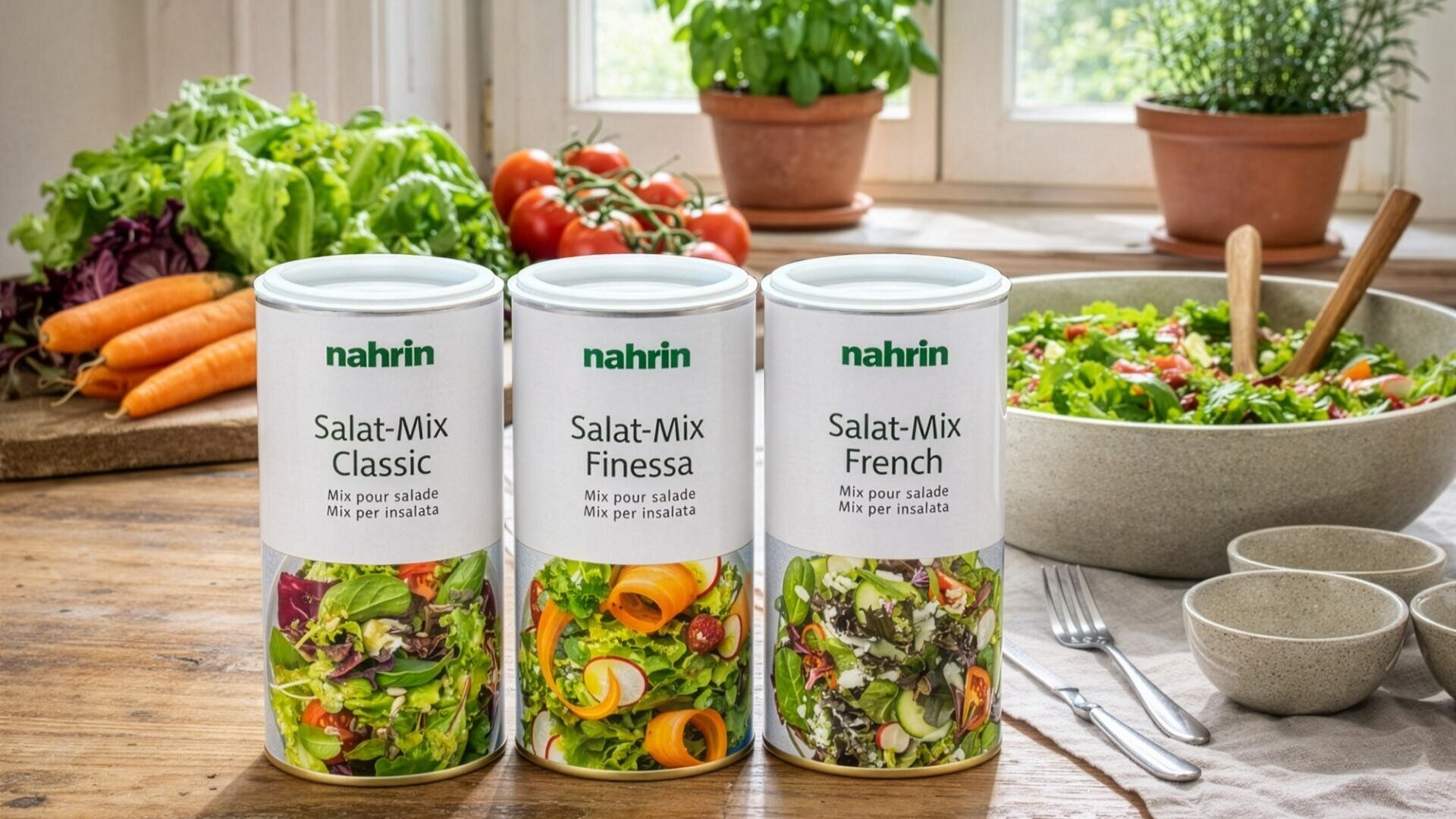 Drei Dosen Nahrin-Salatmischung (Classic, Finessa, French) auf einem Holztisch, umgeben von frischem Gemüse, einer Salatschüssel, Tellern und Besteck, mit einem hellen Fenster und Topfpflanzen im Hintergrund.