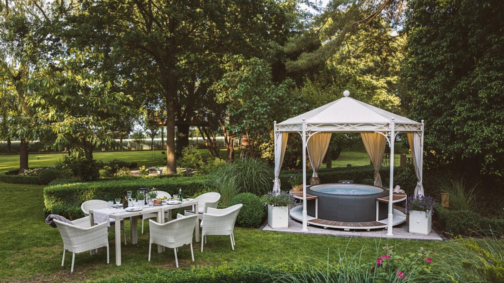 A garden scene with a white outdoor dining table and chairs set for a meal on a lawn, next to a gazebo with a covered hot tub, surrounded by lush greenery and tall trees.