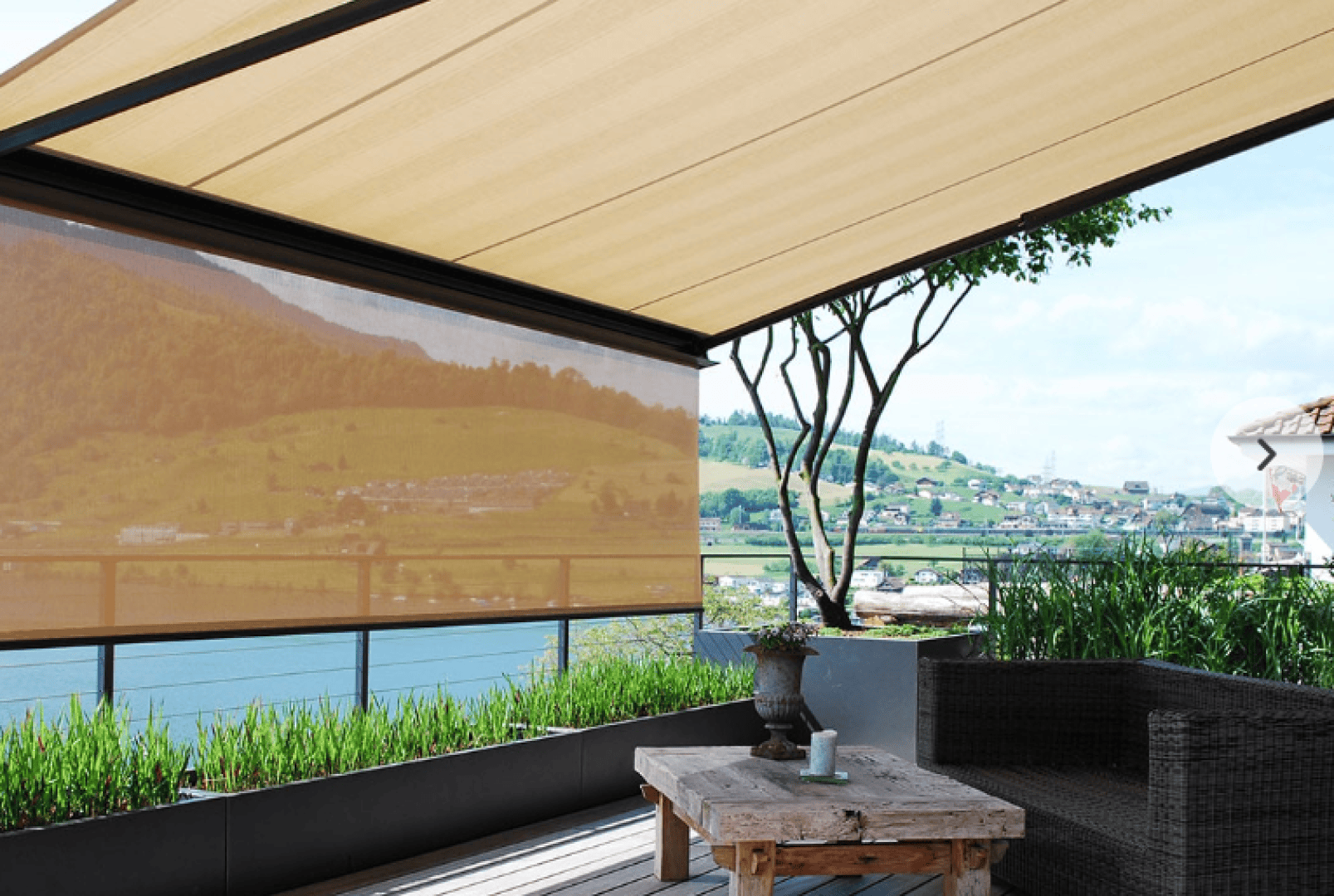 A shaded outdoor patio with a wooden table, wicker sofa, potted plants, and a view of a river, green hills, and a village in the background under a tan retractable awning.