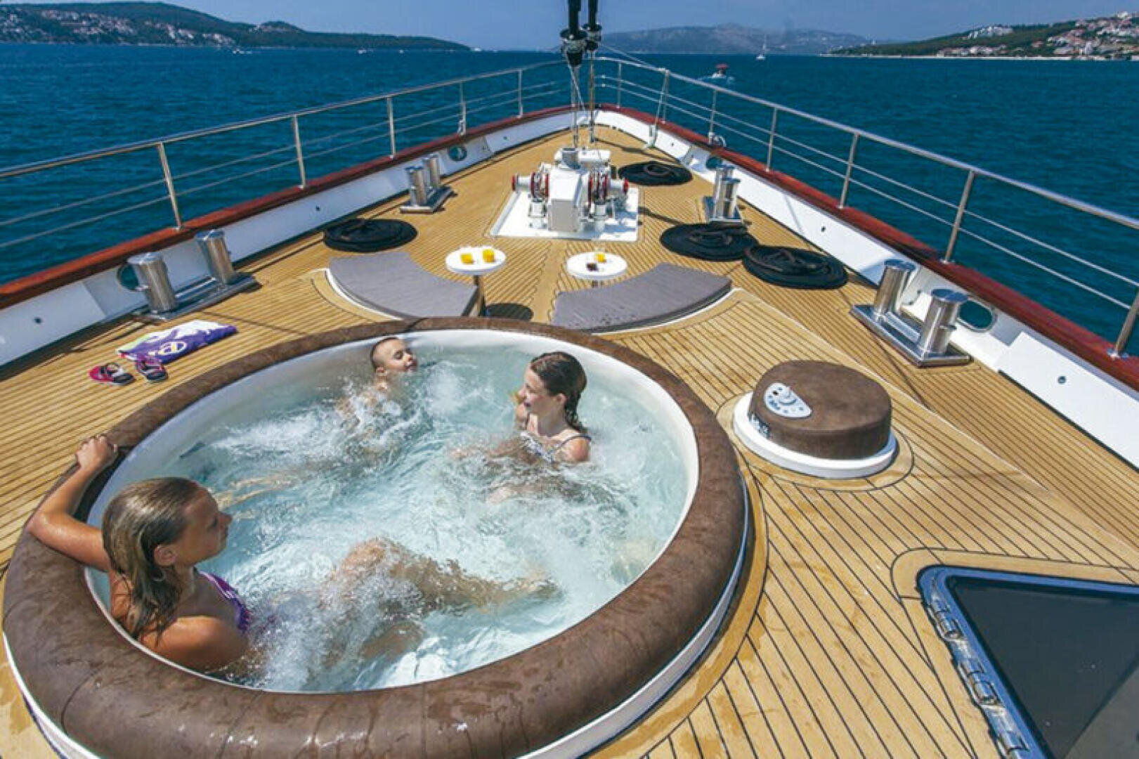 Drei Personen entspannen sich in einem runden Whirlpool auf dem Deck einer Jacht, umgeben von blauem Meer und fernen Hügeln unter einem klaren Himmel. Auf dem Holzdeck befinden sich Sitzgelegenheiten und die Ausrüstung der Jacht.