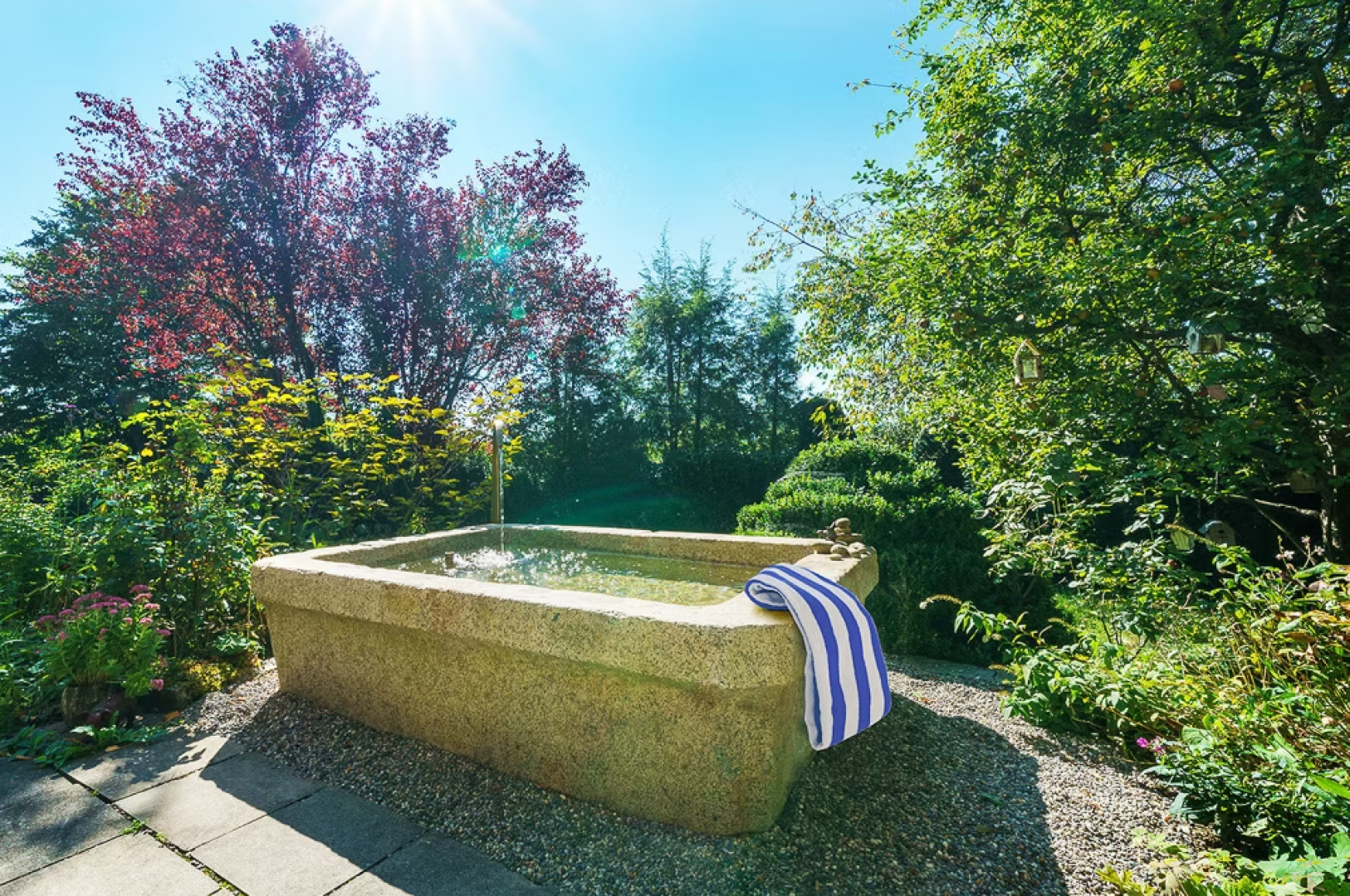 Eine mit Wasser gefüllte Steinbadewanne steht draußen in einem üppigen, sonnendurchfluteten Garten. Ein blau-weiß gestreiftes Handtuch hängt über den Rand. Hohe grüne Bäume und bunt blühende Pflanzen umgeben die Wanne unter einem klaren blauen Himmel.