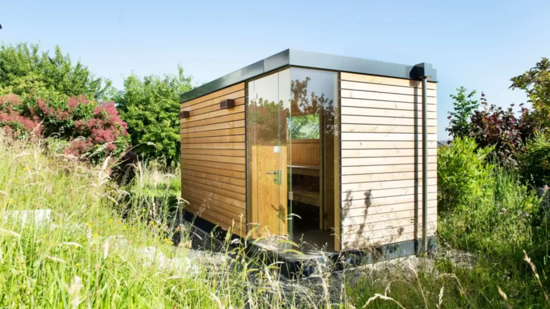 A modern, small wooden hut with vertical cladding stands amidst tall grass and lush greenery under a clear blue sky. The hut has large glass doors and minimalist design features.
