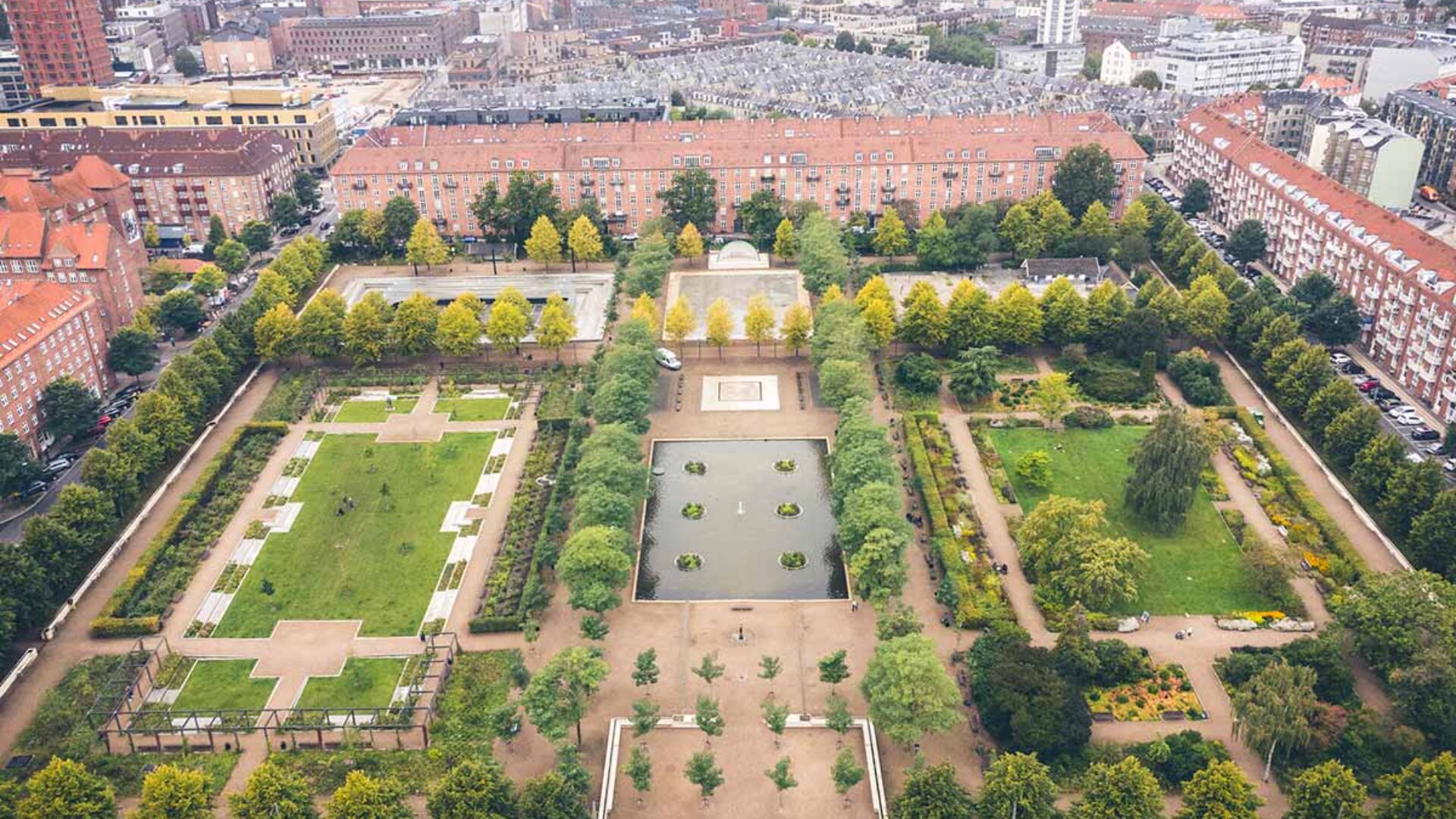 Luftaufnahme eines symmetrischen Stadtparks mit rechteckigen Rasenflächen, von Bäumen gesäumten Wegen, einem zentralen Teich und umliegenden Gebäuden mit roten Dächern in einer Stadtlandschaft.