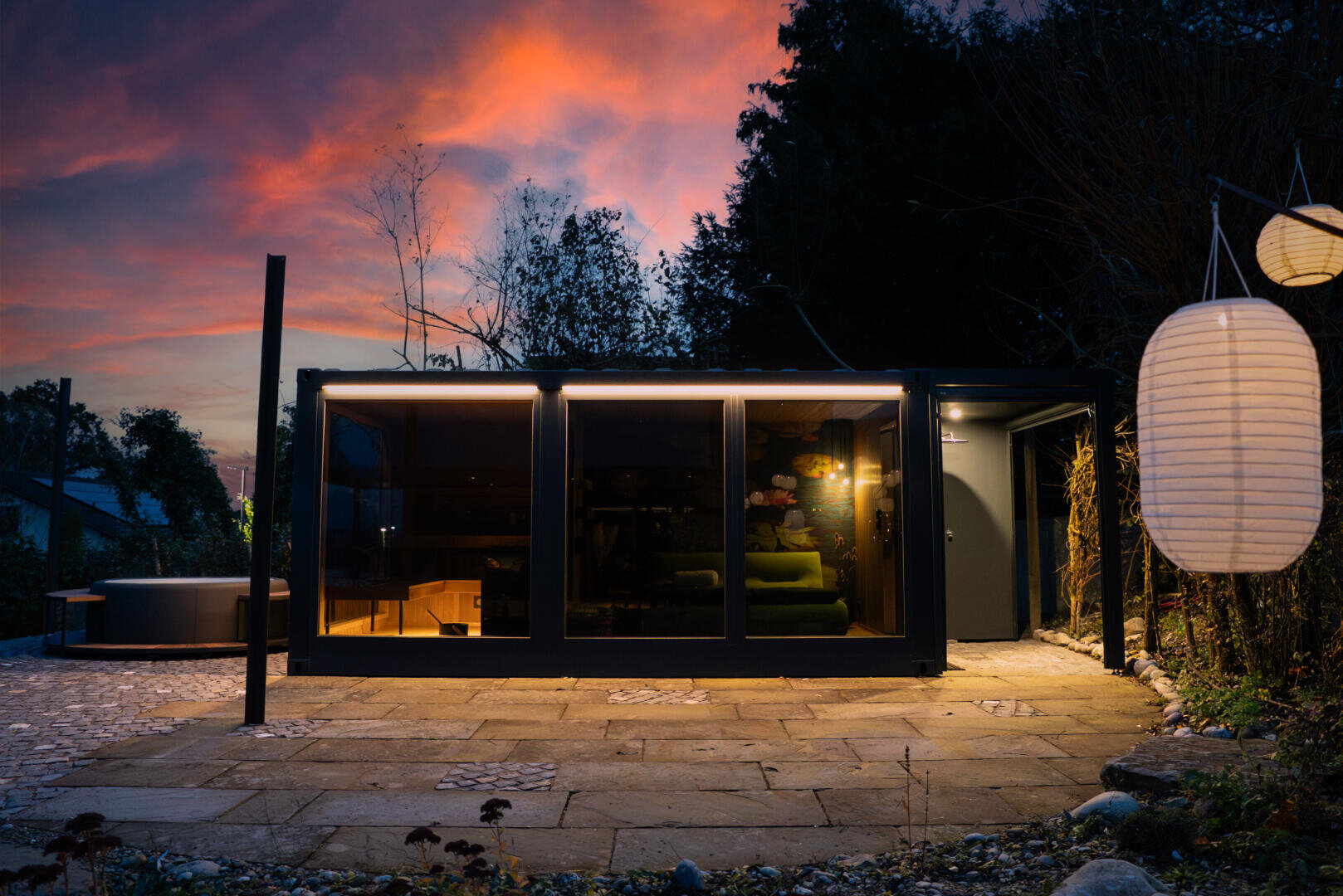Eine moderne, gläserne Hütte wird in der Abenddämmerung beleuchtet, mit warmem Licht im Inneren und einem leuchtenden Sonnenuntergangshimmel im Hintergrund. Rechts hängen Papierlaternen, und eine Steinterrasse führt zum Eingang. Die Szene ist von Bäumen umgeben.