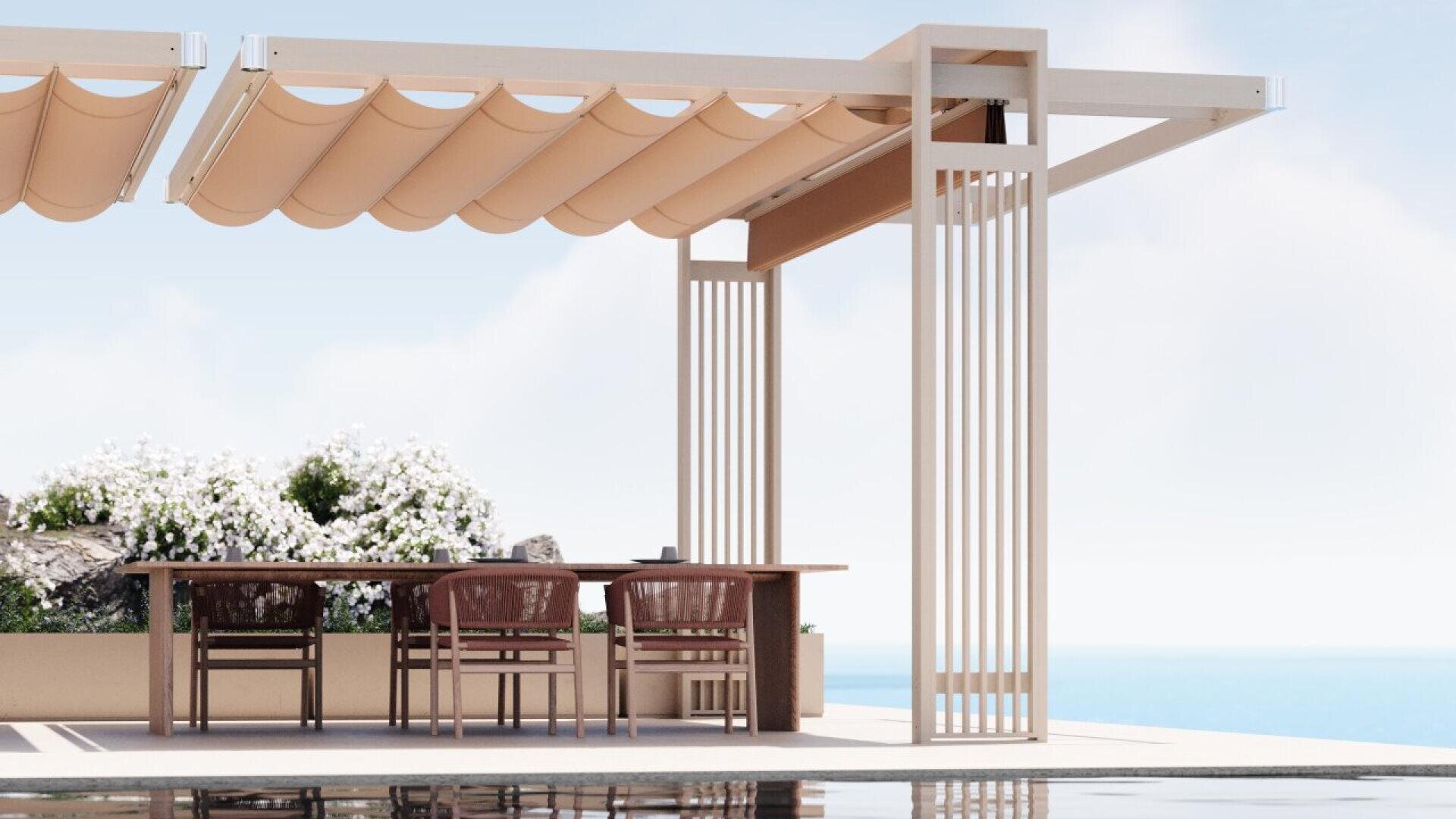 A modern pergola with a beige retractable canopy shades a wooden dining set with four chairs. White flowers line the back, and the scene overlooks a calm blue sea under a clear sky.
