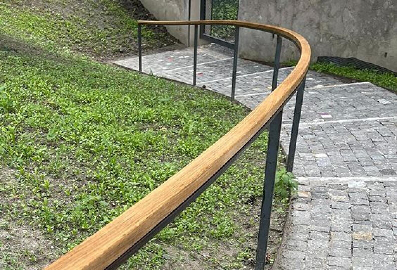 Ein sanft geschwungener hölzerner Handlauf folgt einem mit Steinen gepflasterten Weg, der auf der einen Seite von grünem Gras und auf der anderen Seite von einer Betonmauer begrenzt wird. Der Weg führt zu einer Glastür im Hintergrund.
