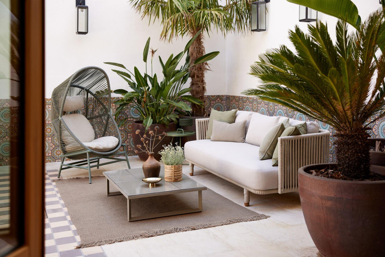 Stilvolle Außenterrasse mit weißem Sofa, rundem Loungesessel, niedrigem Couchtisch, grünen Topfpflanzen, gefliesten Wänden und einem neutralen Teppich, der eine entspannte und gemütliche Atmosphäre schafft.