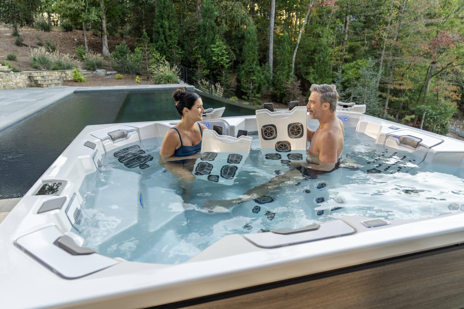 A man and a woman relax and chat in a modern outdoor hot tub near a pool, surrounded by trees and greenery in the background.