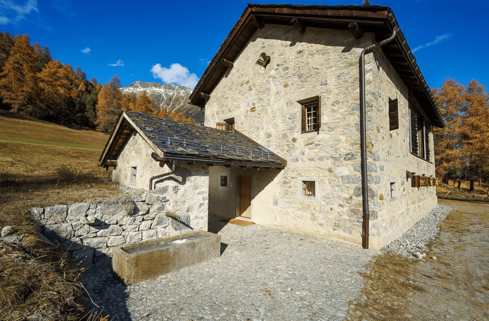 A rustic stone house with a small extension stands on a gravel path, surrounded by autumnal trees and distant snow-capped mountains under a bright blue sky.