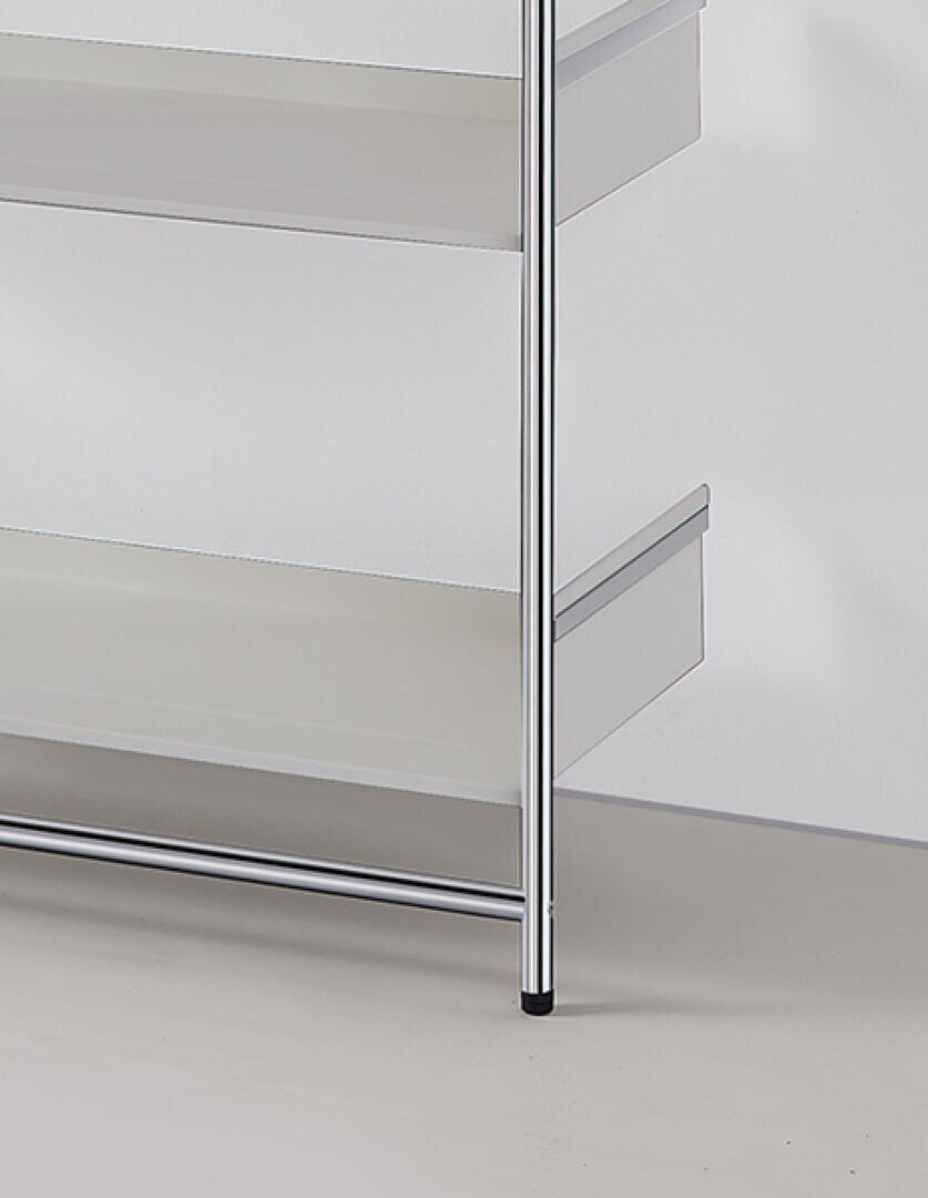 Minimalistisches Metallregal mit verchromtem Rahmen und weißen Einlegeböden vor einer schlichten weißen Wand auf einem hellen Boden. Nur der untere rechte Teil des Regals ist auf dem Bild zu sehen.