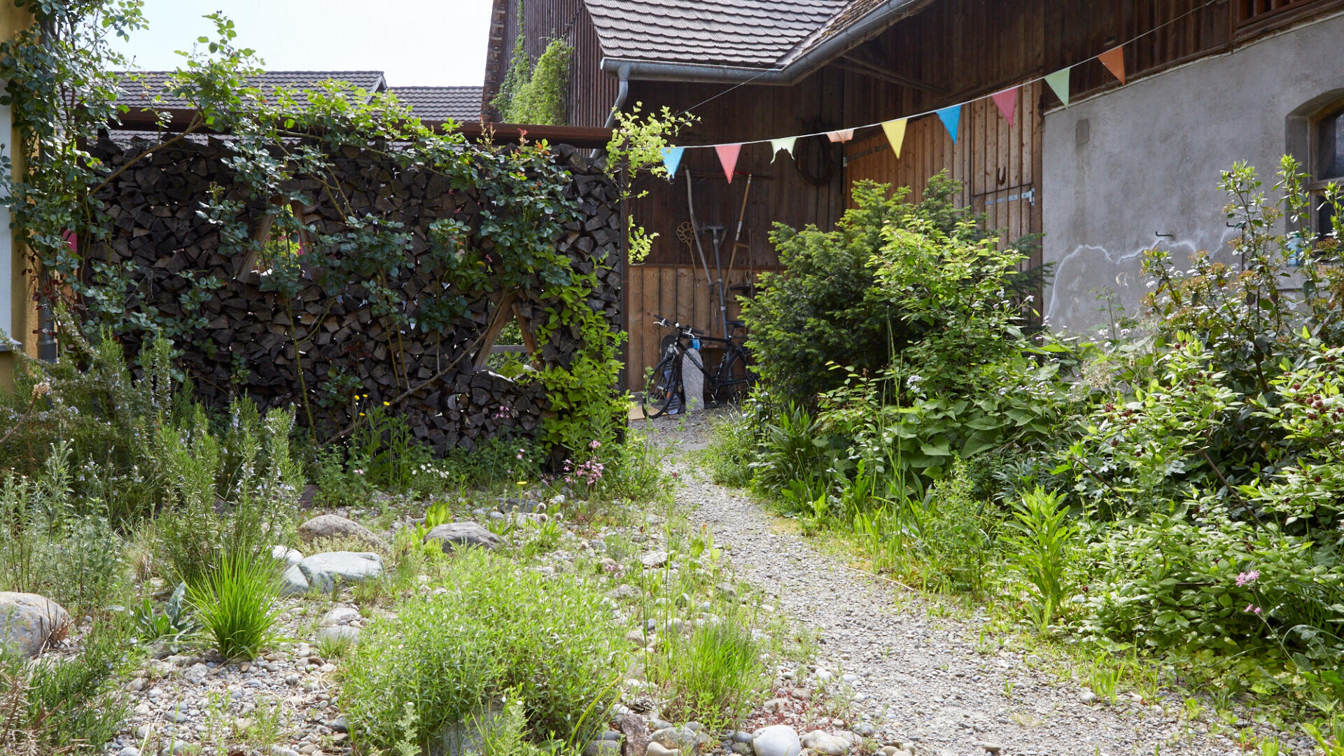 Ein mit Grünpflanzen und Sträuchern gesäumter Kiesweg führt zu Holzgebäuden. Darüber hängen bunte Wimpelfahnen, und auf der linken Seite ist ein Stapel Brennholz zu sehen. Im Hintergrund in der Mitte ist ein Fahrrad geparkt.