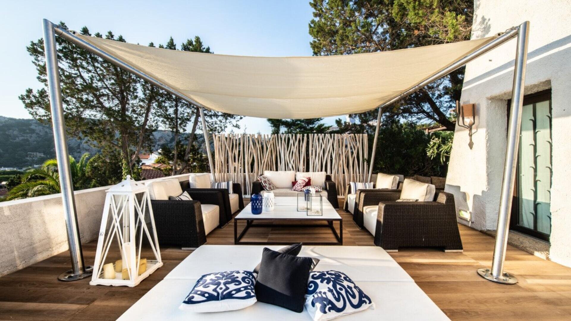 Modern outdoor patio with a canopy, wicker furniture, cushions, a coffee table with lanterns, and decorative pillows on a white rug. Surrounded by greenery and a partial white wall.